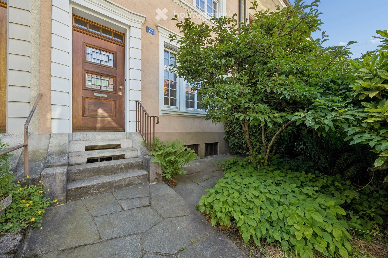 Front entrance of a two-story house with a wooden door, steps, and lush greenery surrounding the pathway.