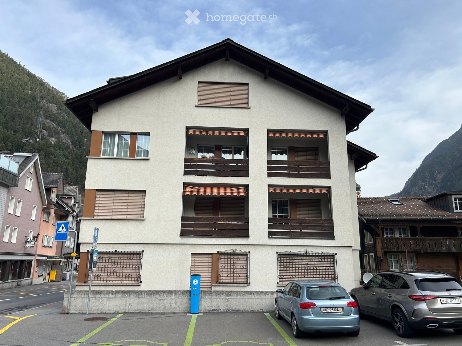 A two-story residential building with balconies, located in a mountainous area with parked cars in front.