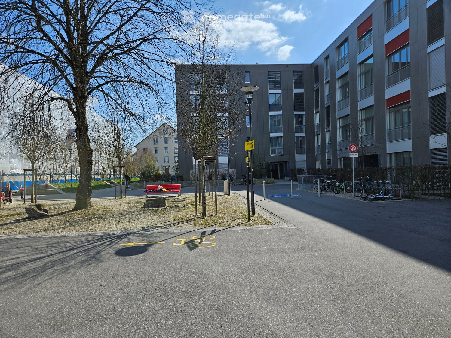 Courtyard area with modern apartment buildings, a red bench, bicycles, and a playground in the background.