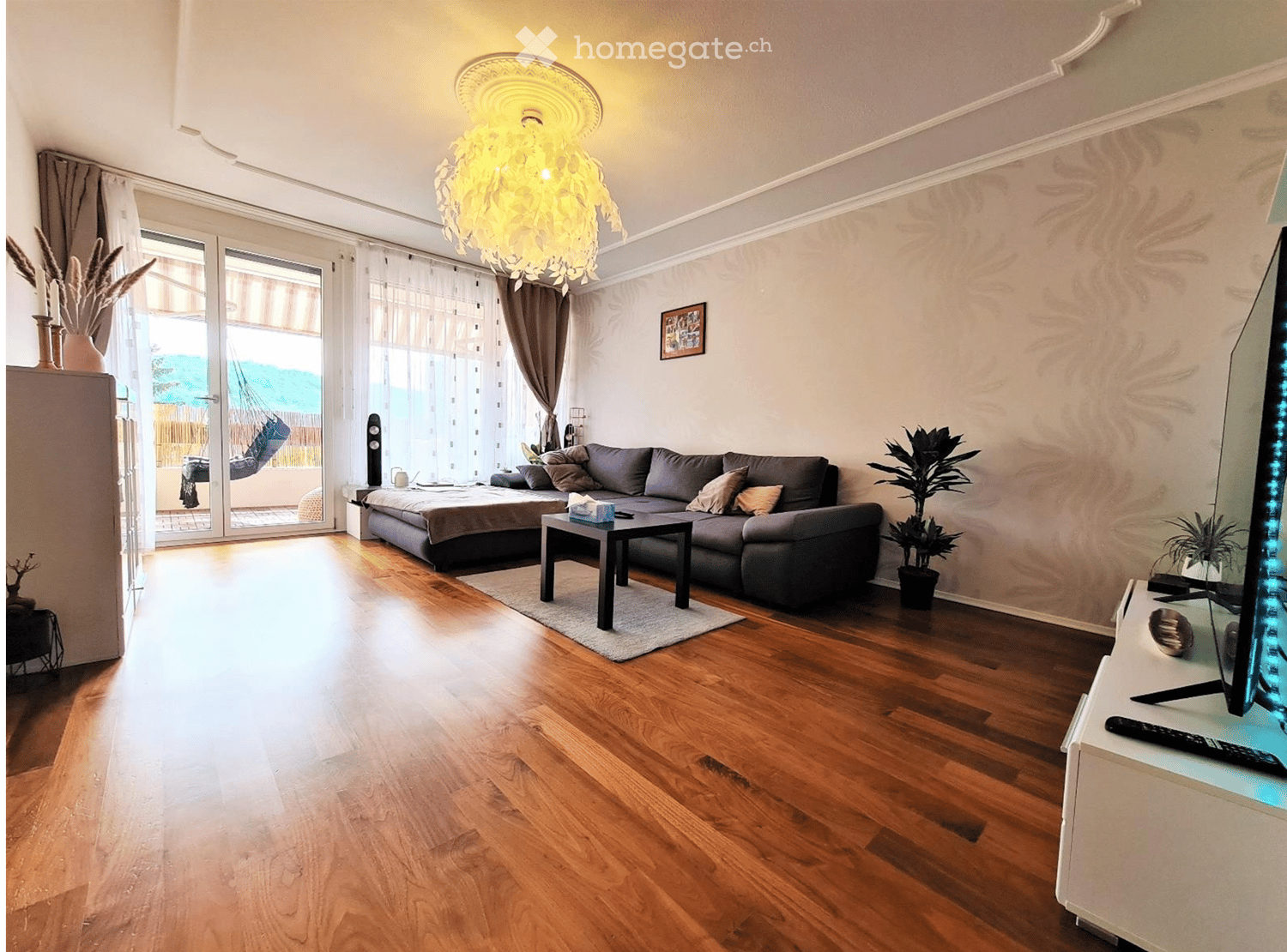 Modern living room with wooden flooring, grey sectional sofa, coffee table, TV, decorative plants, and a chandelier.
