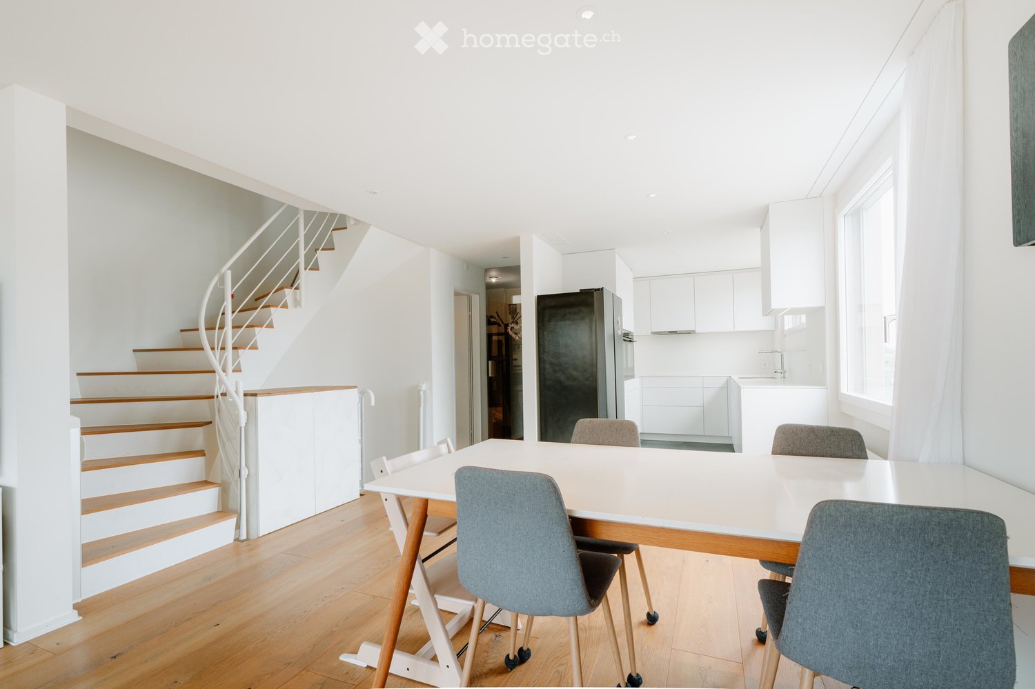 Bright, open-plan dining area with a modern wooden dining table and chairs, leading to a compact kitchen with white cabinets and a refrigerator. A spiral staircase is visible in the background.