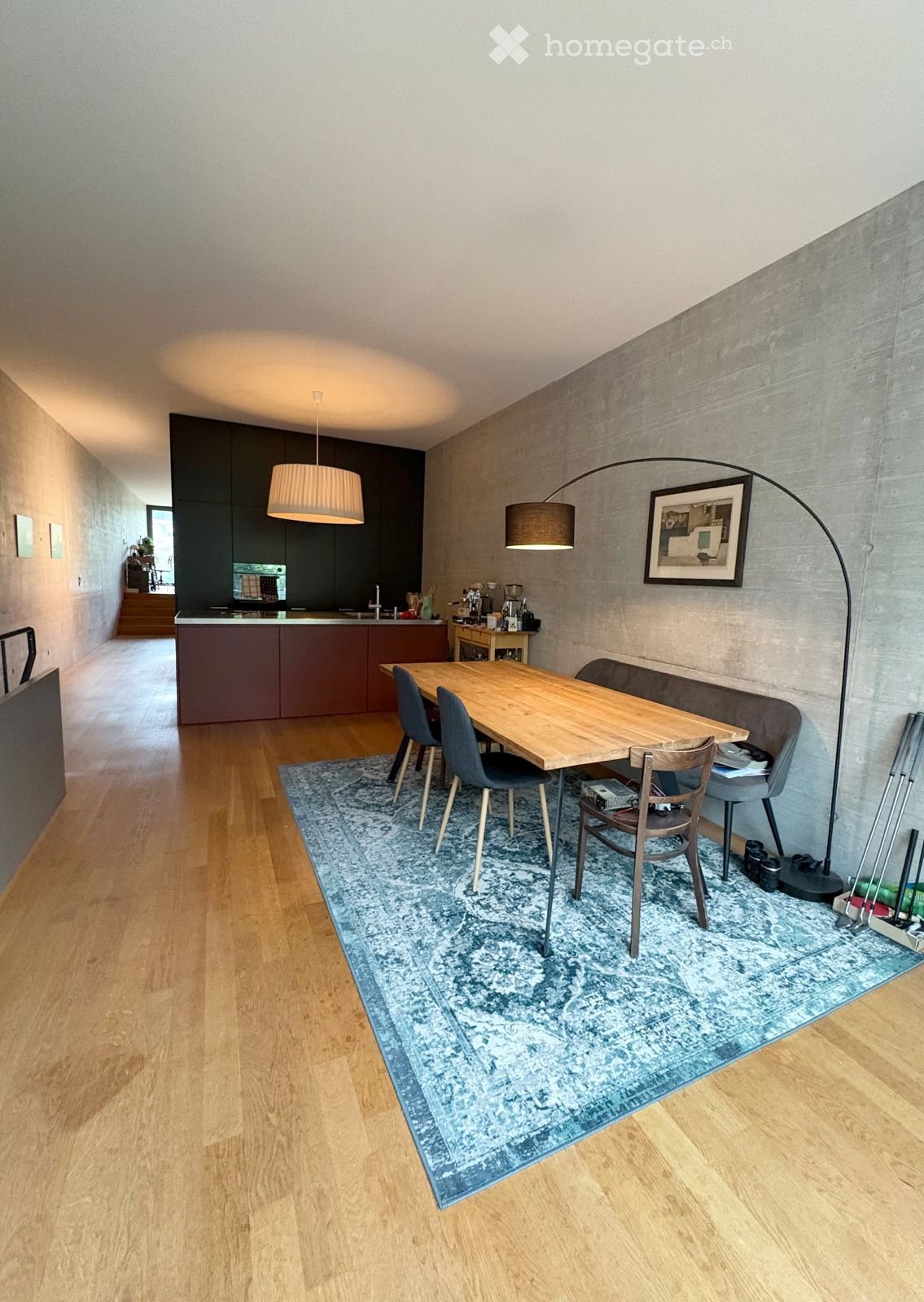 Dining area with a wooden table, four chairs, a sofa, a pendant light, a floor lamp, a framed picture, and a kitchen area with cabinets and a coffee machine.