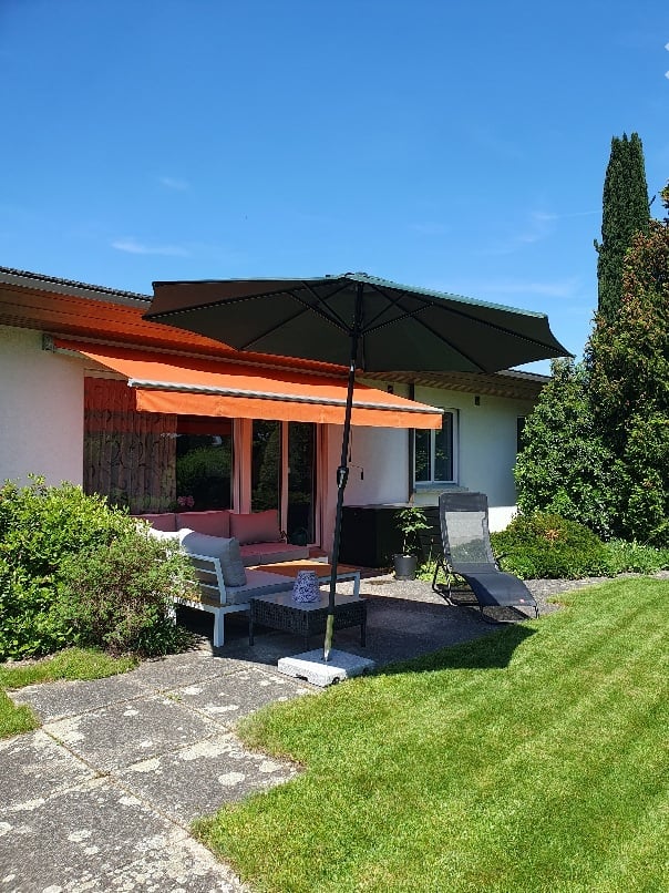 A well-maintained garden area with a patio, outdoor furniture including a sofa, chairs, and a large umbrella, surrounded by greenery and a house with a covered entrance.
