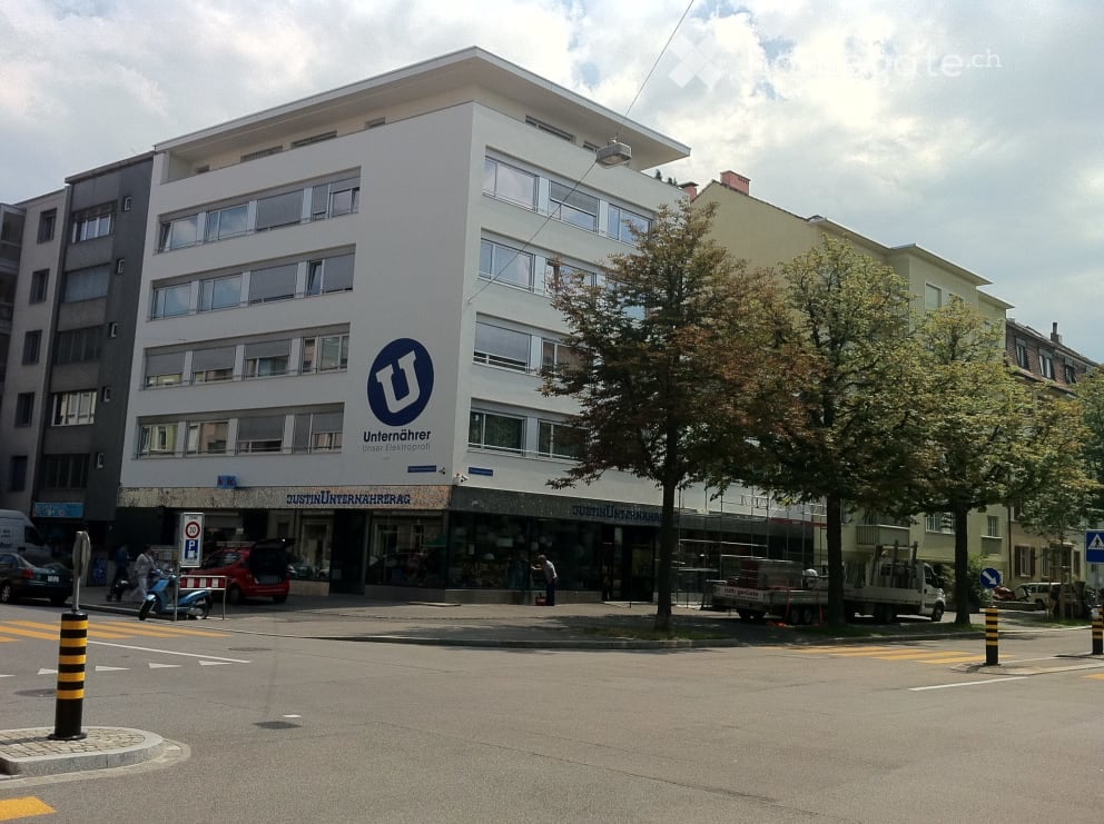 Modern multi-story building with large windows and a sign for "Unternehmer Union" on the facade. The building is situated on a street corner with trees and traffic signs visible.