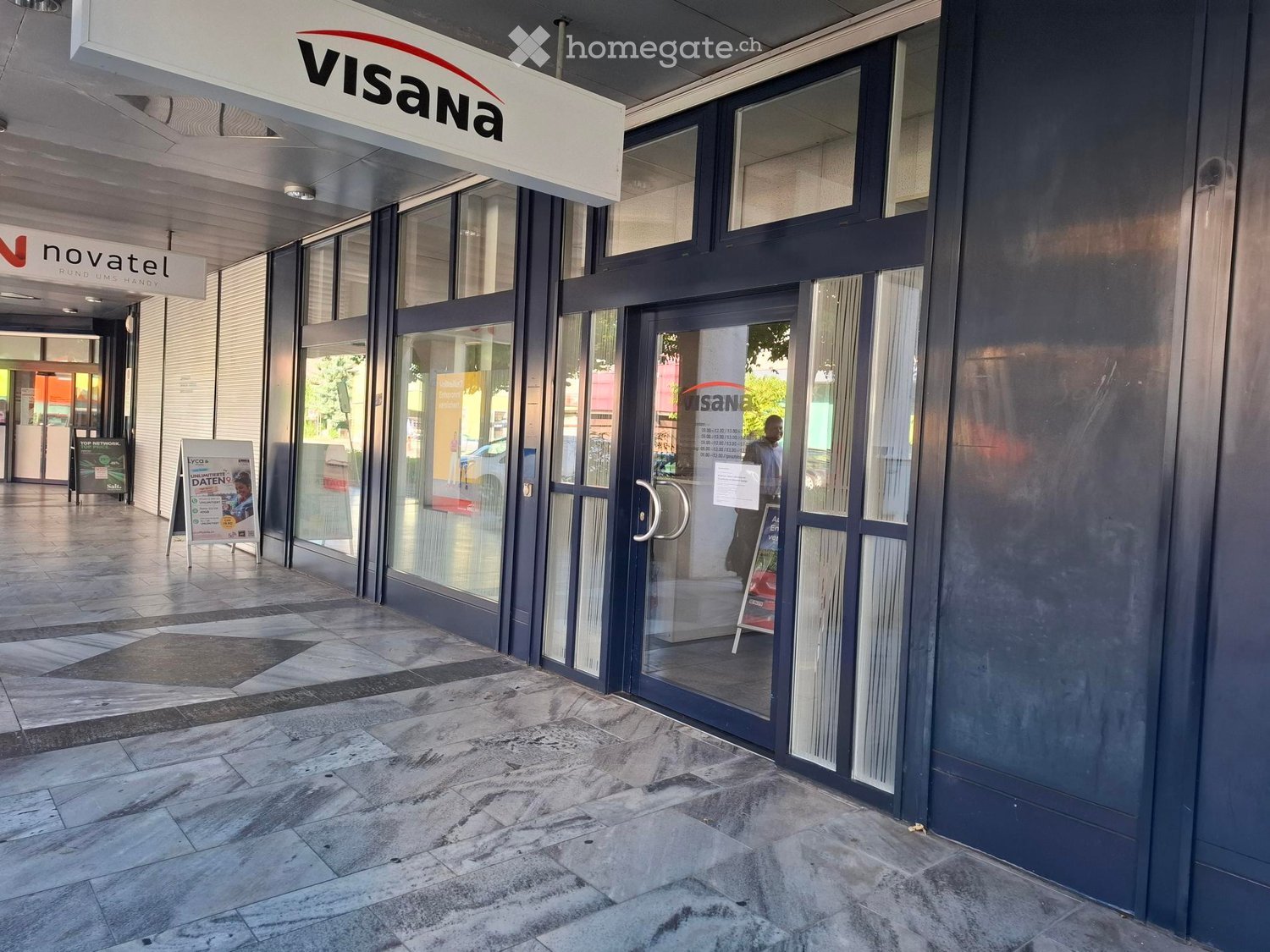 Entrance to a commercial building with glass doors, signage for "Visana" and "Novatel" above, tiled floor, and a few informational signs nearby.