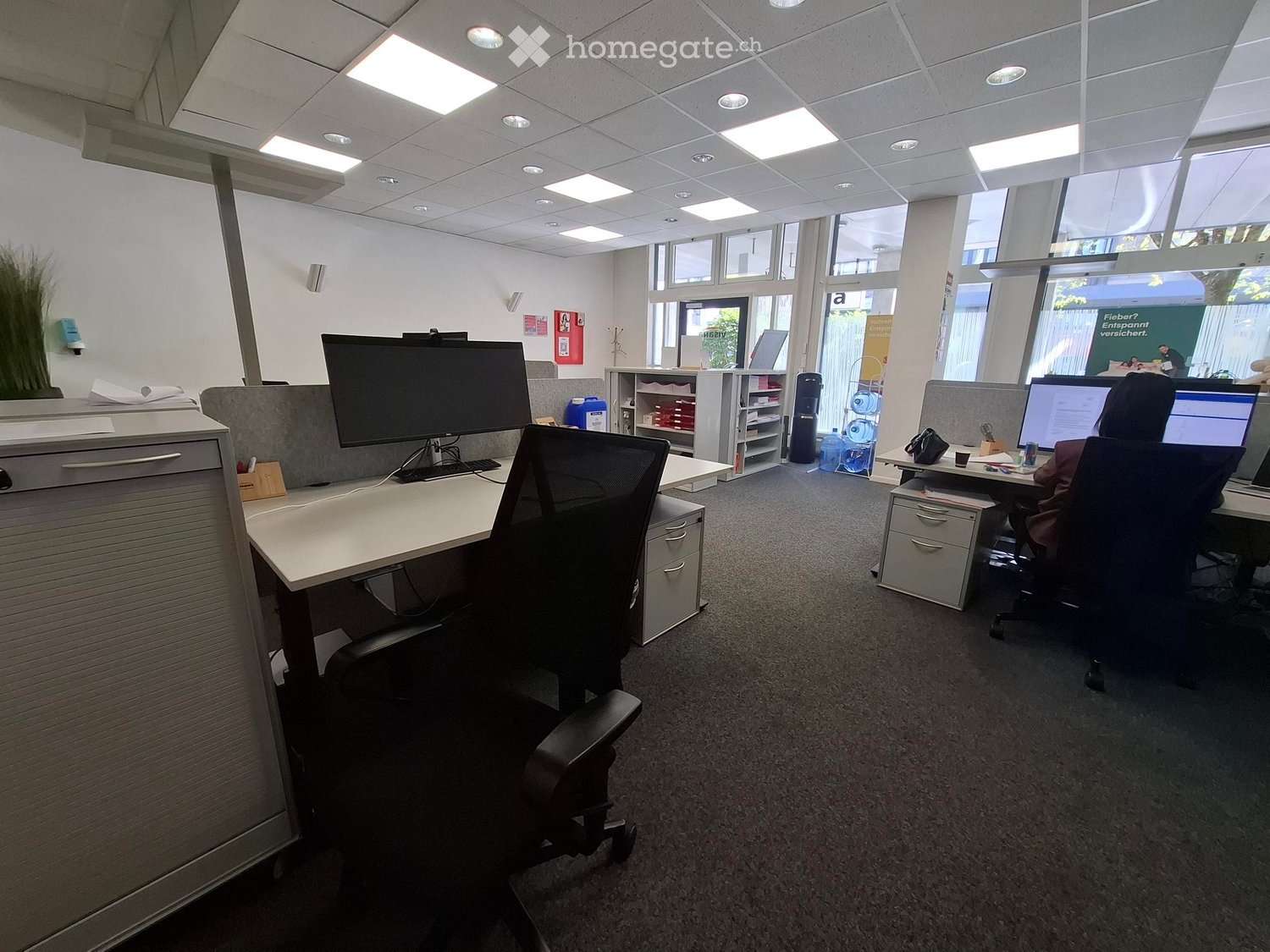 Modern office space with desks, chairs, computers, and a filing cabinet. Large windows provide natural light, and the room is equipped with a plant for decoration.
