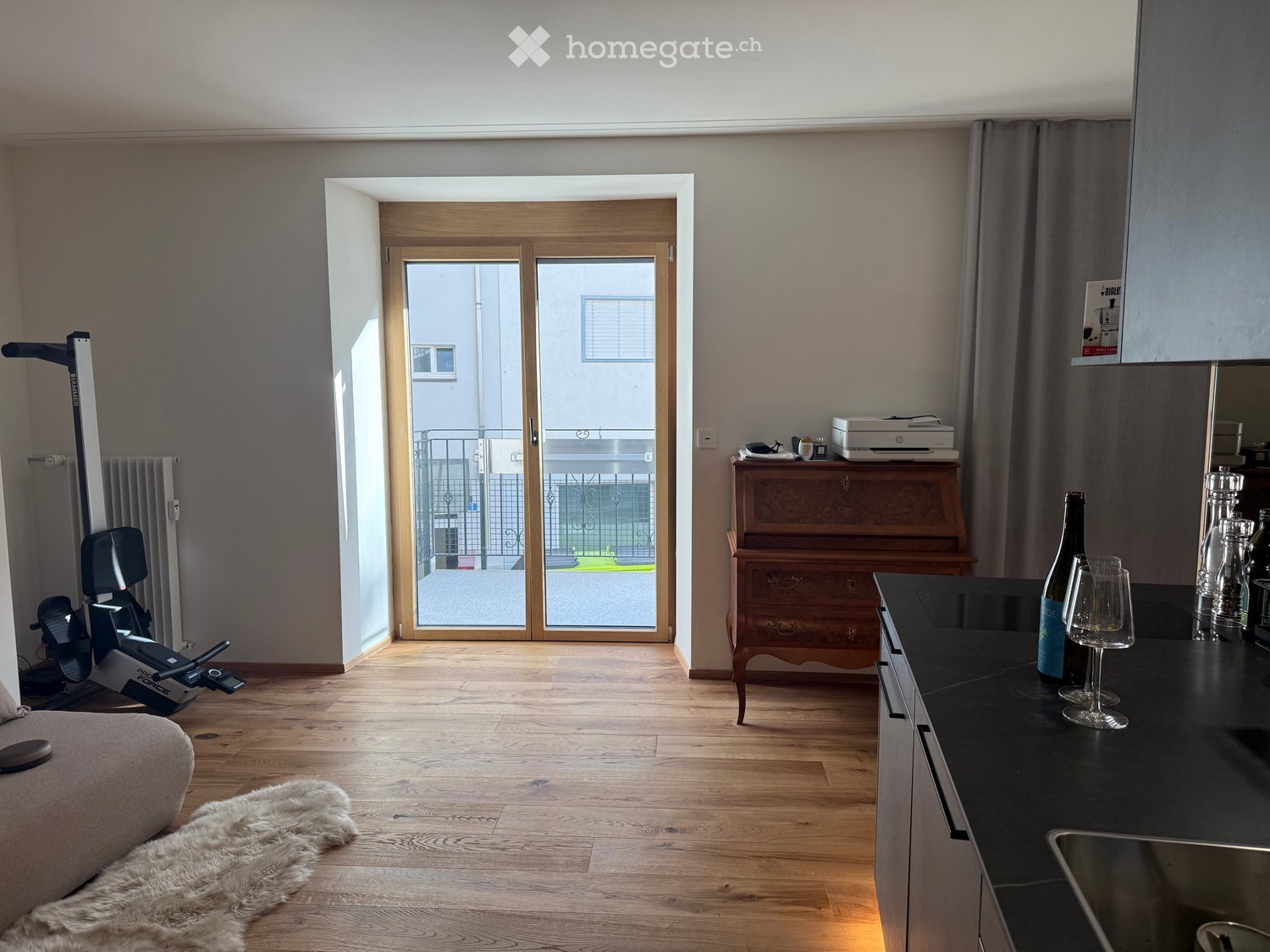 A modern, well-lit living space with wooden flooring, a sofa, an exercise bike, a piano, and a kitchen area with a countertop, wine bottle, and wine glasses.