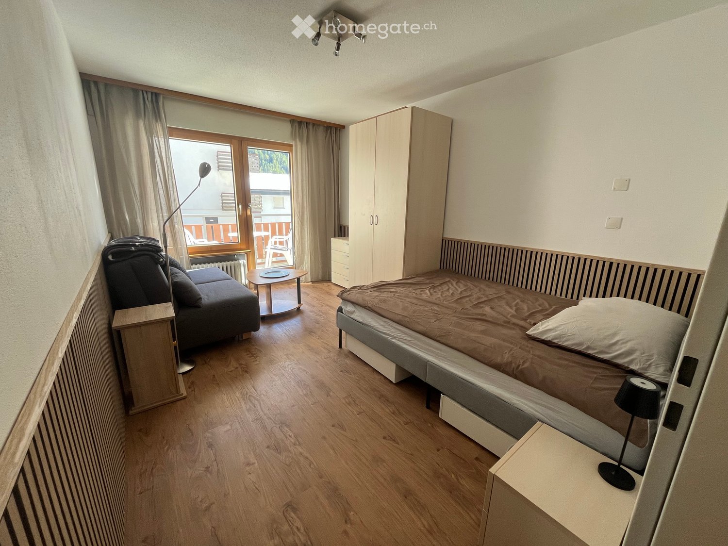 A room with wooden flooring, single bed, sofa, table, cabinet, radiator, and view through a large window.