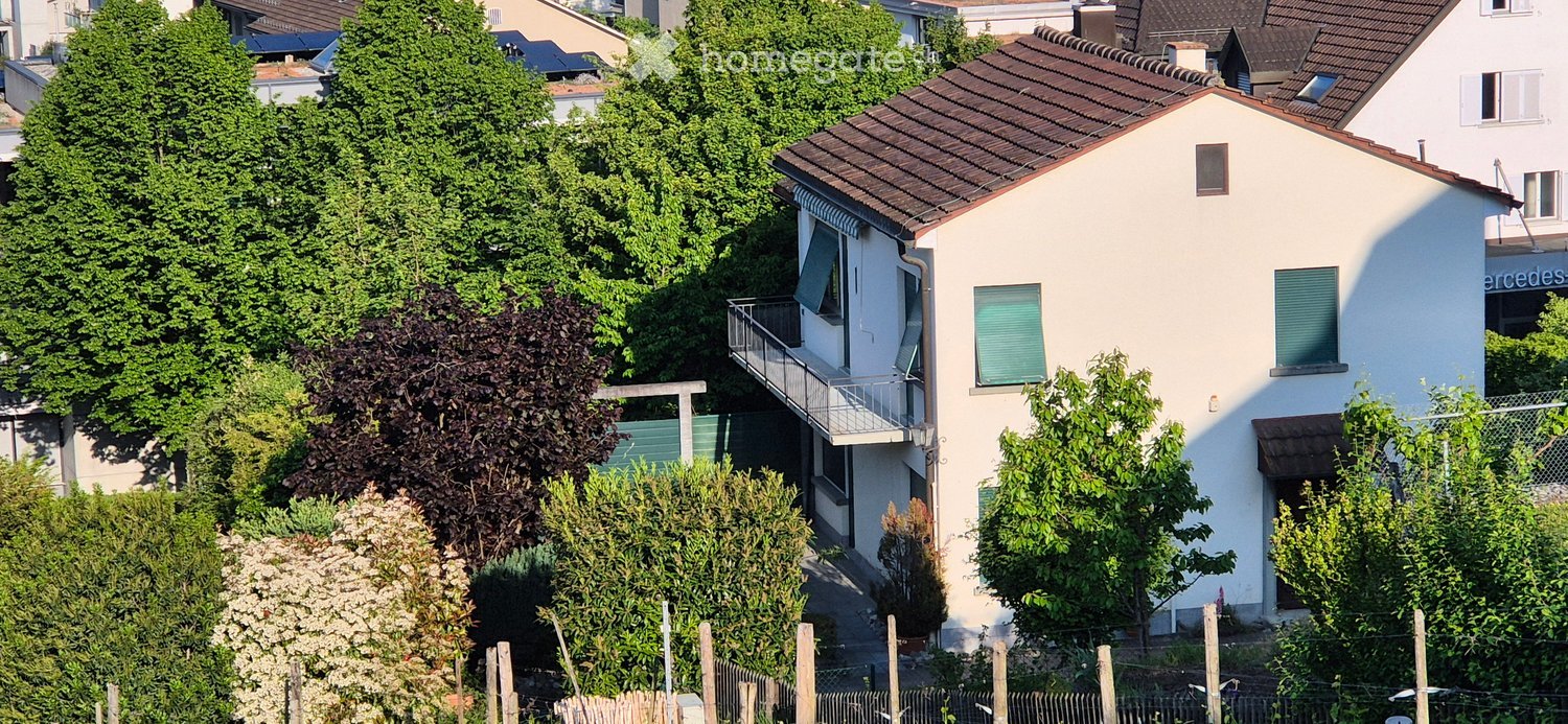 Two-story house with white exterior, green shutters, and a balcony on the upper floor, surrounded by trees and greenery.