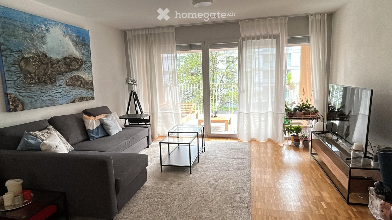 Modern living room with a grey sectional sofa, coffee table, side tables, TV, floor lamp, and large windows with white curtains. A painting hangs on the wall, and a small table with a chair is visible near the balcony.