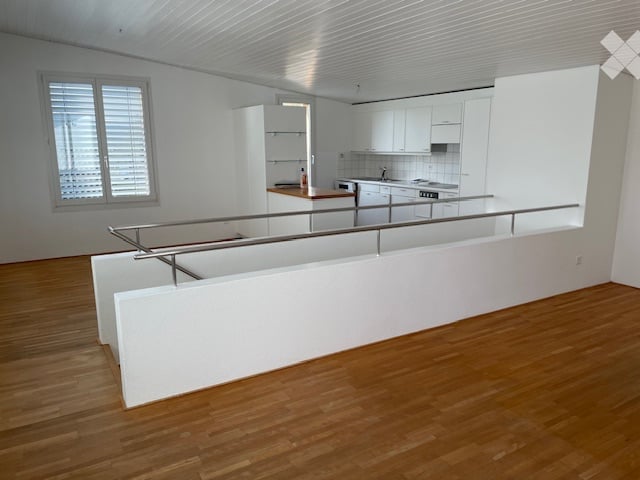 Open-plan space with modern white kitchen cabinets, wooden flooring, and a white partition wall. The area includes a kitchen section with built-in appliances and a dining area with a long white bench and metal railing.