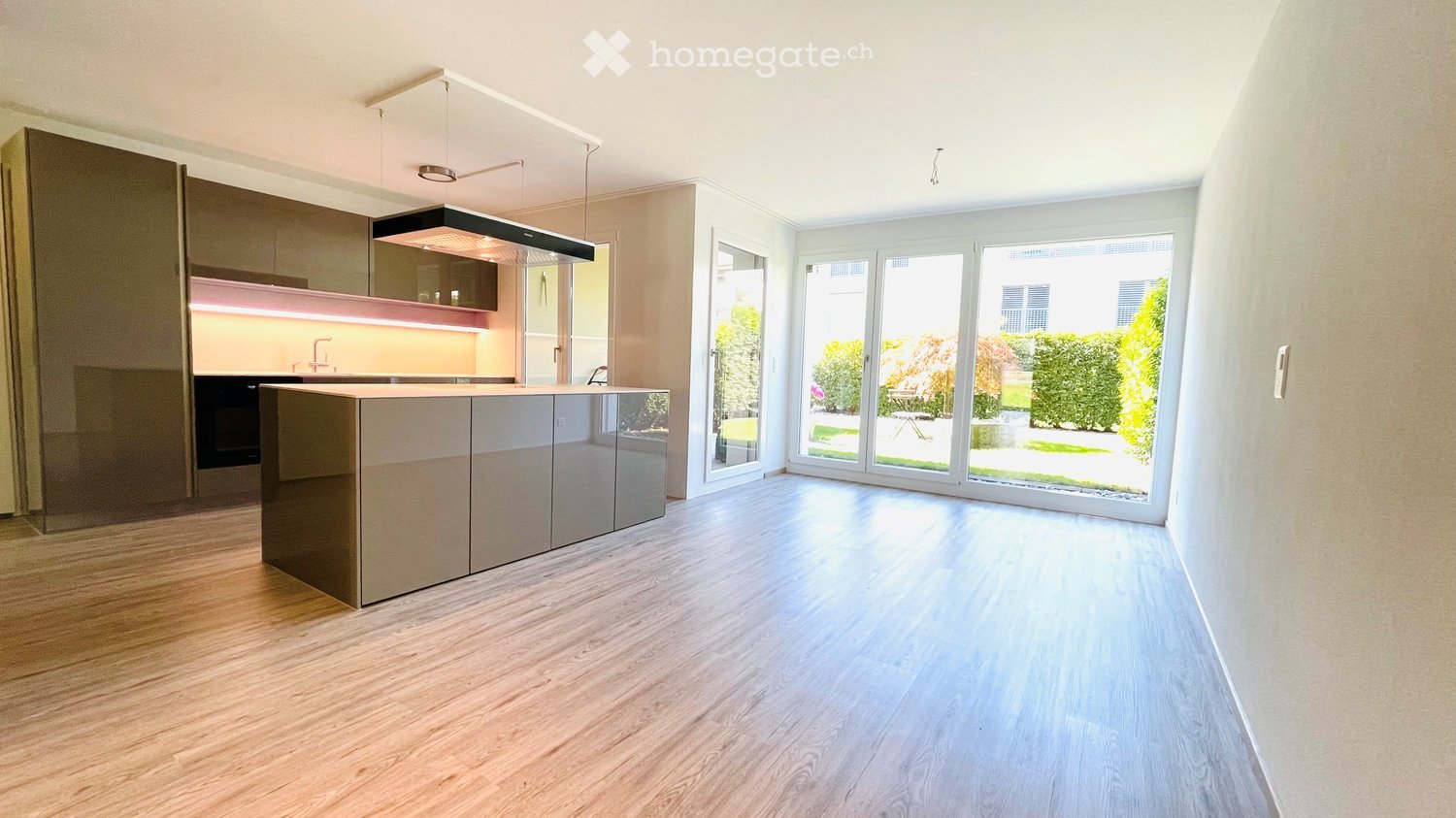 Modern kitchen with light wood flooring, sleek cabinetry, and a central island. Large windows provide natural light and a view of the garden.