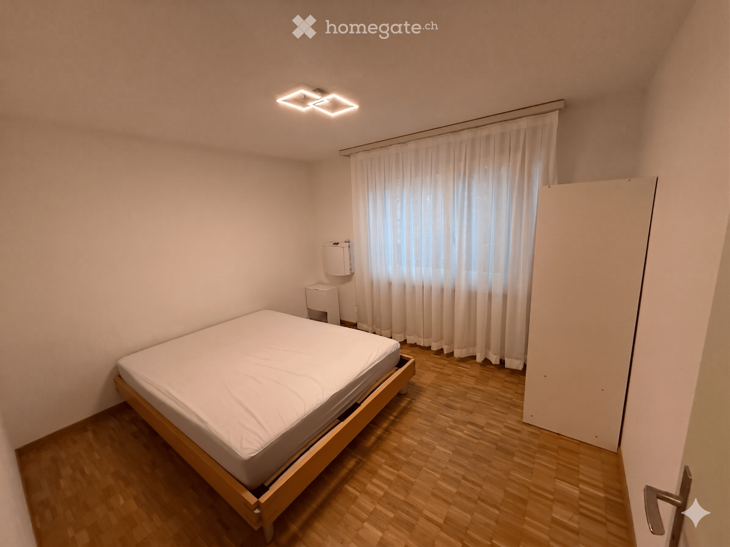 Minimalist bedroom with a wooden bed frame, white bedding, a window with white curtains, and a radiator on the wall.