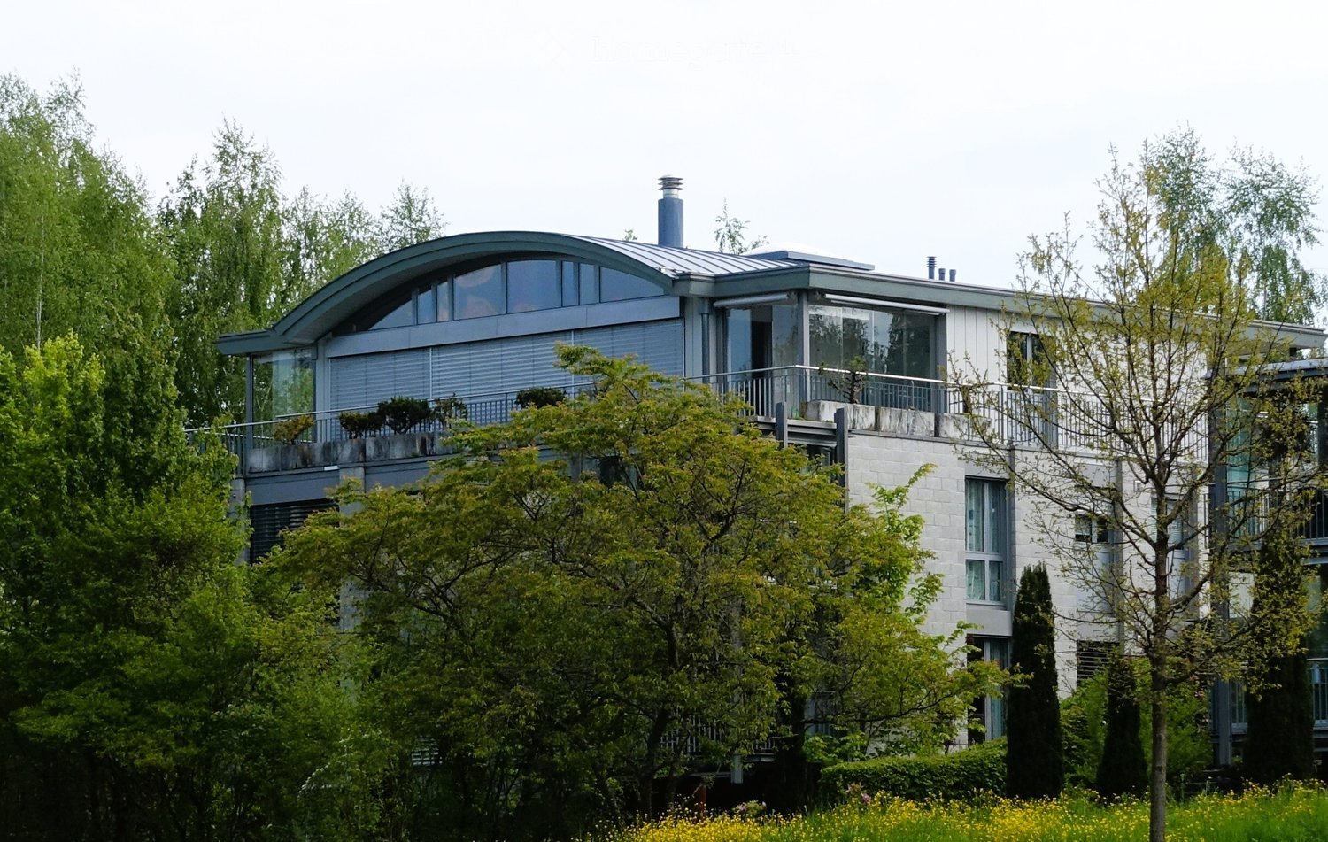 Modern two-story house with a curved roof, large windows, and a balcony, surrounded by lush greenery and trees.