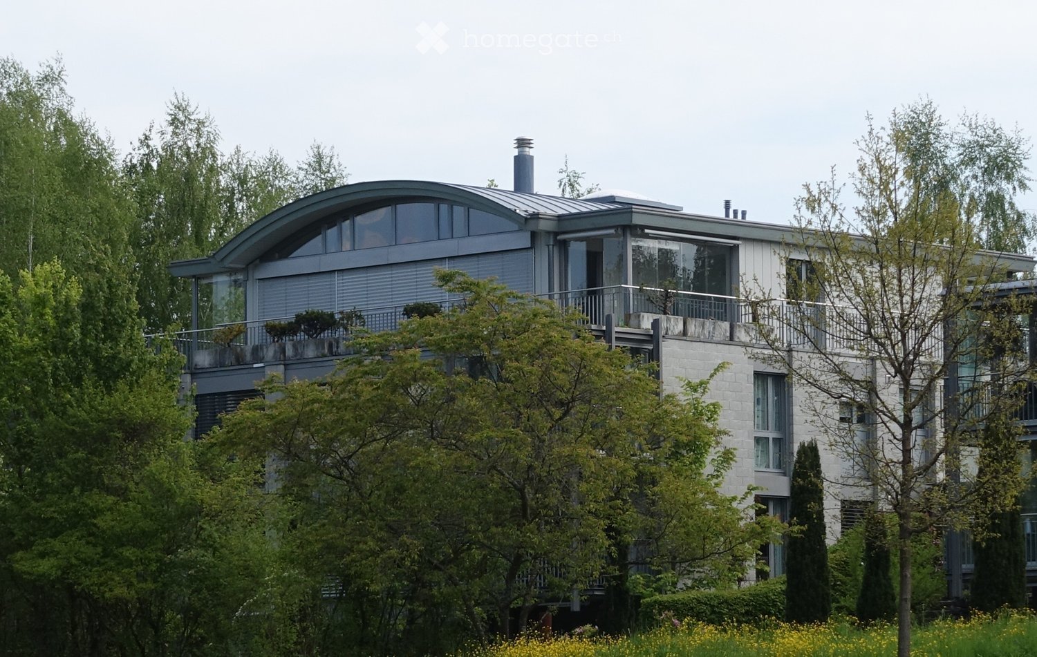Modern two-story house with a curved roof, large windows, and a balcony, surrounded by lush greenery and trees.