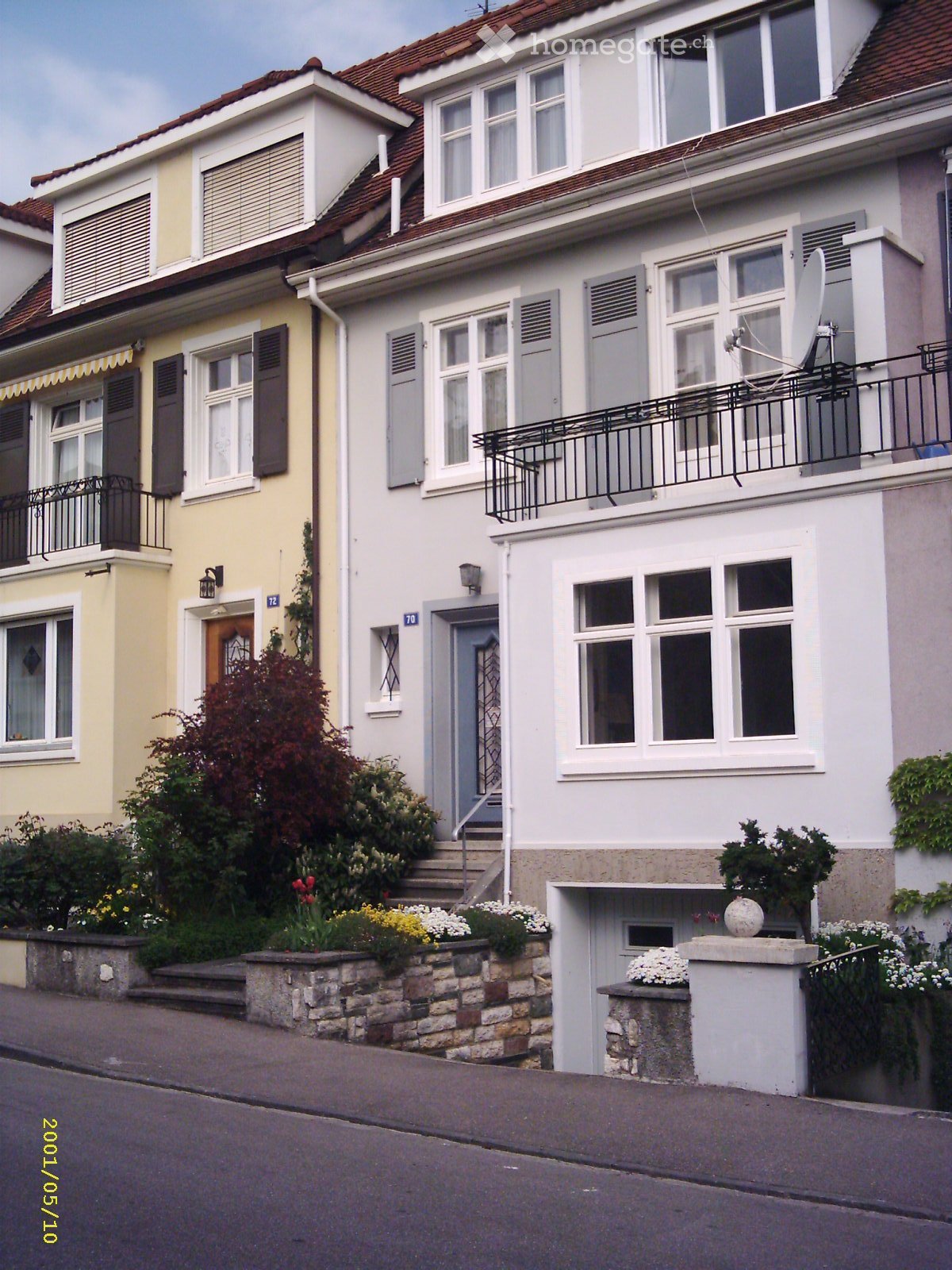 Two-story residential house with white and beige exterior, multiple windows with shutters, balconies, and a small garden area with flowers and shrubs.