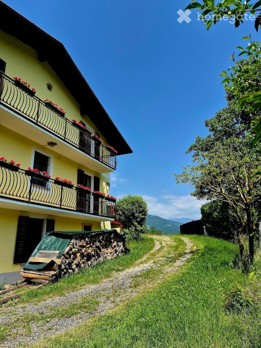 Two-story yellow house with balconies, flower boxes, and a stone woodpile near the entrance. A gravel path leads to the house, surrounded by greenery and a scenic mountain view.