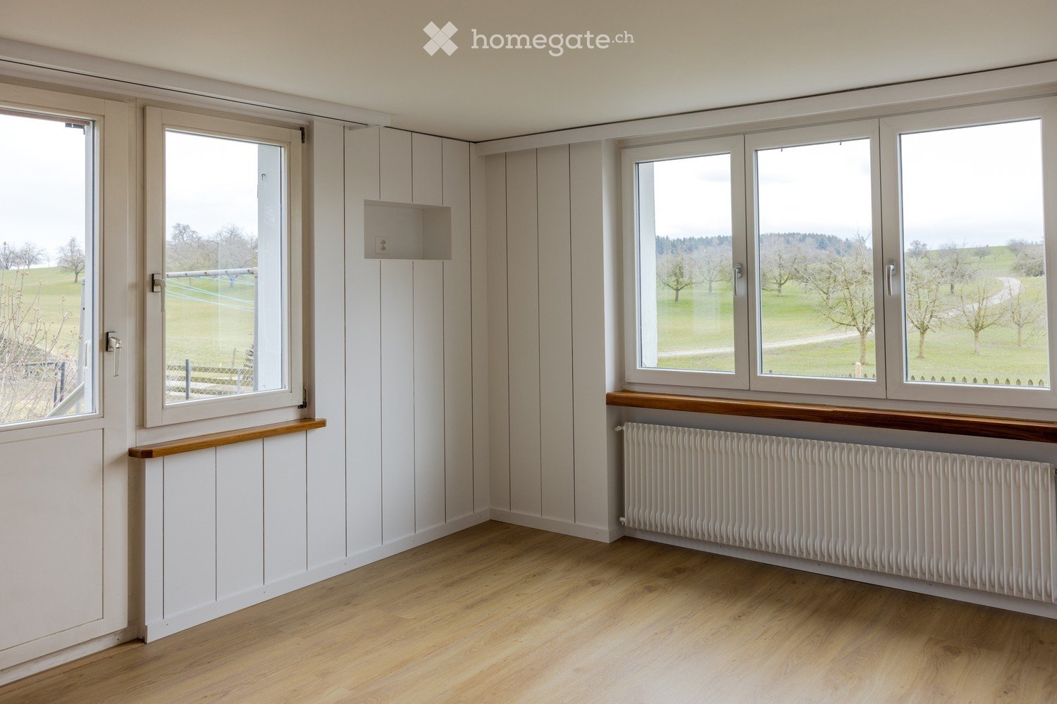 Wooden floor, white wooden panels on walls, glass doors, windows with brown frames, radiator below windows