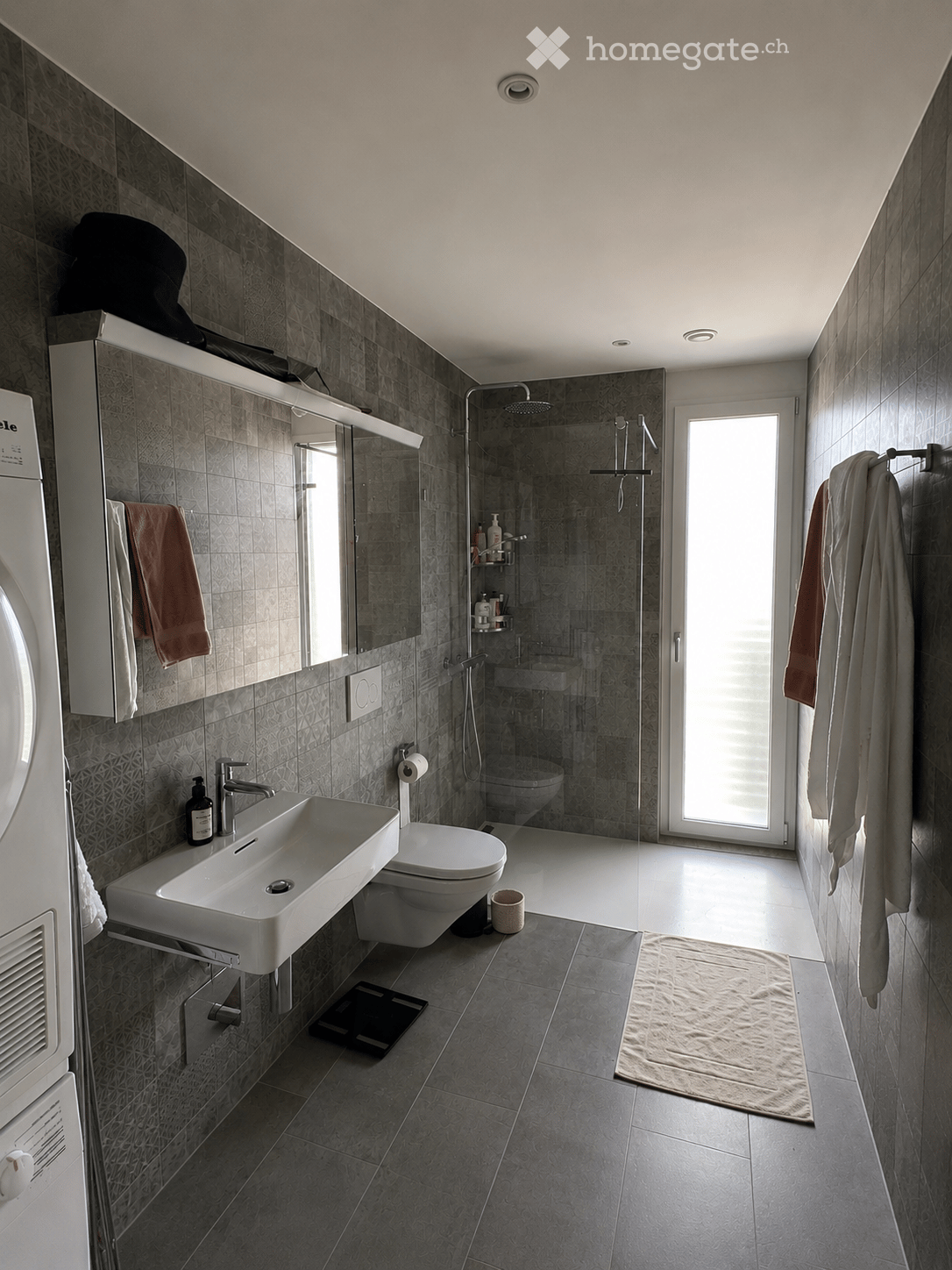 Modern bathroom with grey tiles, a white sink, toilet, shower, and a large mirror. Towels are hanging on the wall, and a small rug is placed on the floor.
