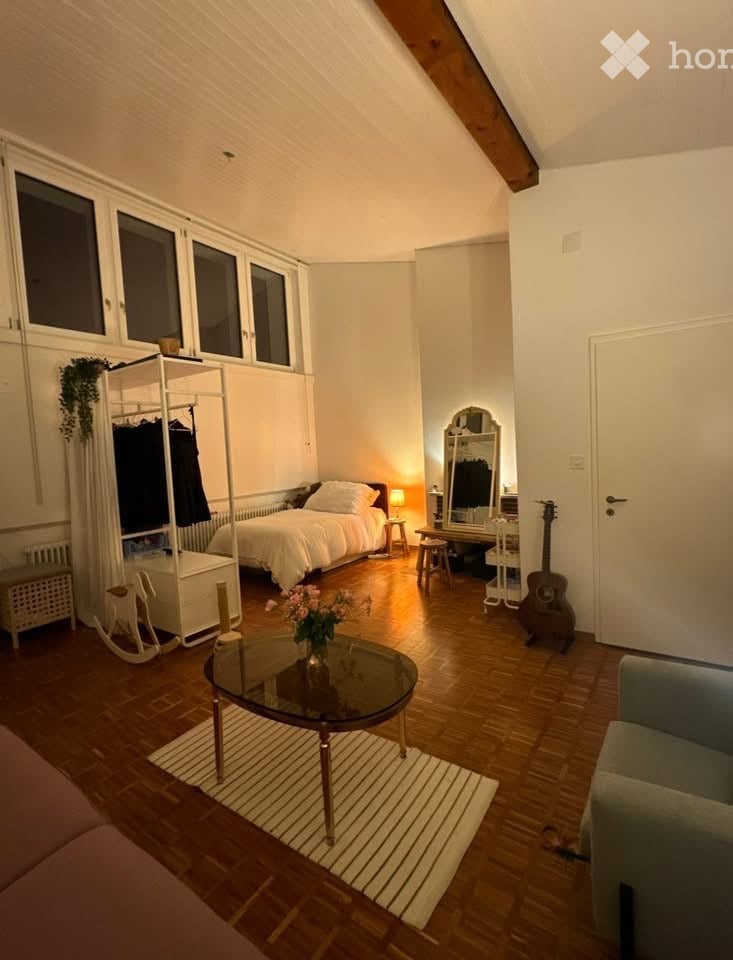 Cozy attic bedroom with wooden floors, large windows, a bed, a sofa, a coffee table, a guitar, a mirror, and a wardrobe.
