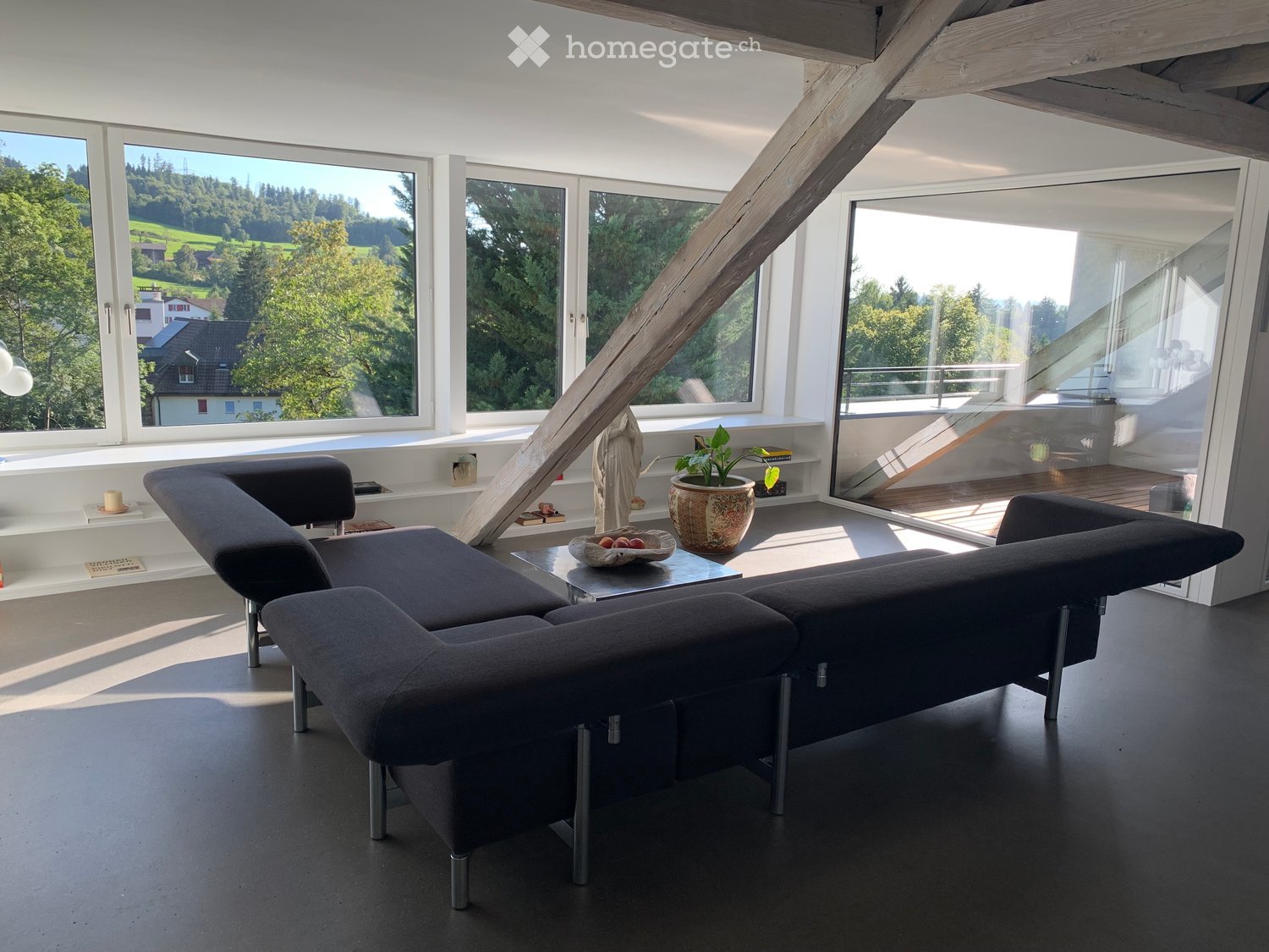 Modern living room with large windows offering a scenic view, featuring a grey sectional sofa, a glass coffee table, a potted plant, and decorative items.