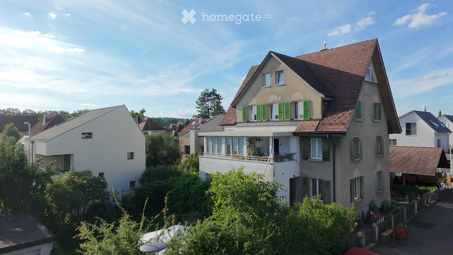 A multi-story residential house with a red-tiled roof, green shutters, and a balcony on the upper floor. The house is surrounded by greenery and trees, with neighboring houses visible in the background.