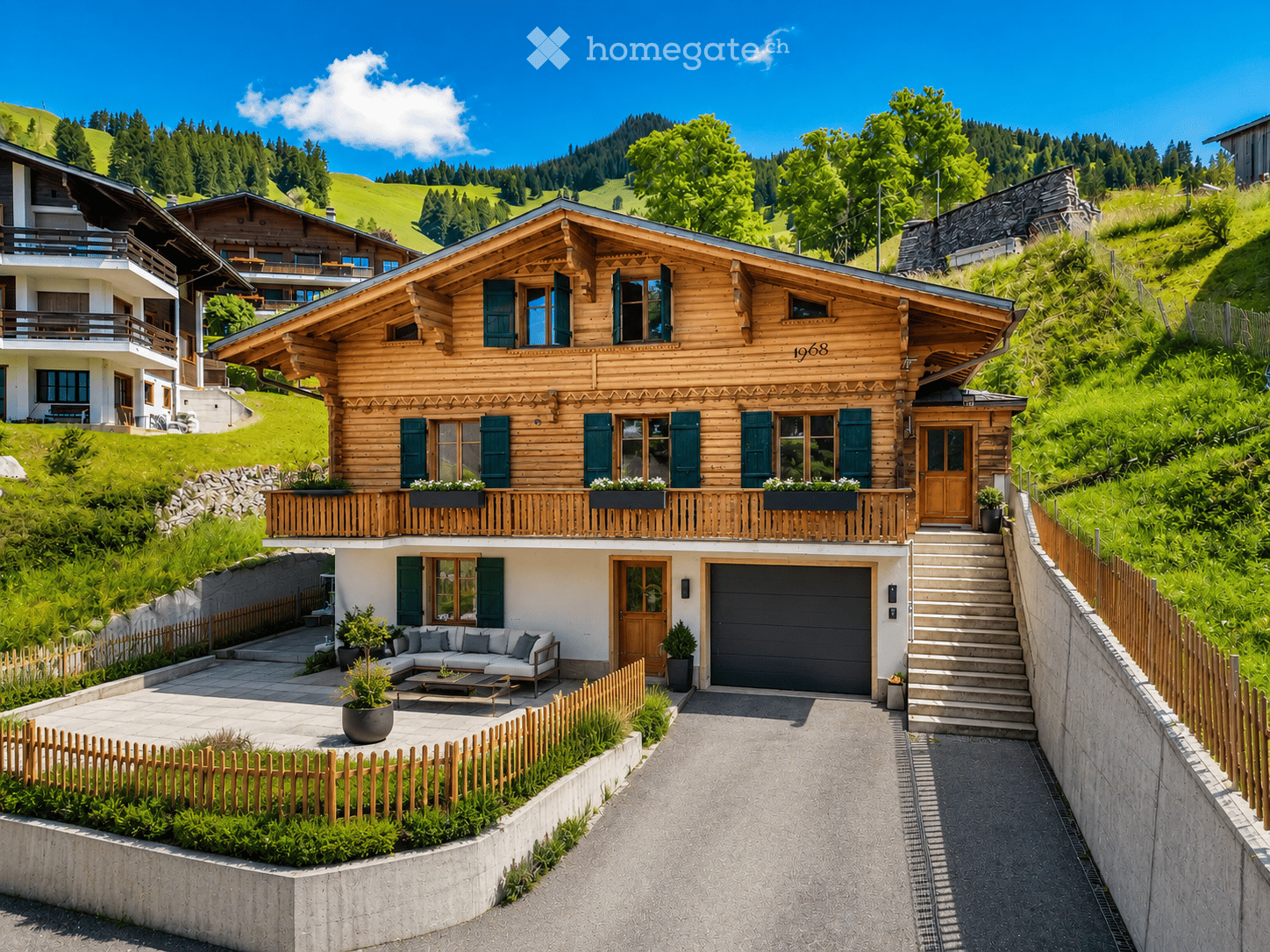 A two-story wooden house with green shutters, a balcony, and a garage, situated in a lush, green landscape with a raised ground floor and a view of the surrounding hills.