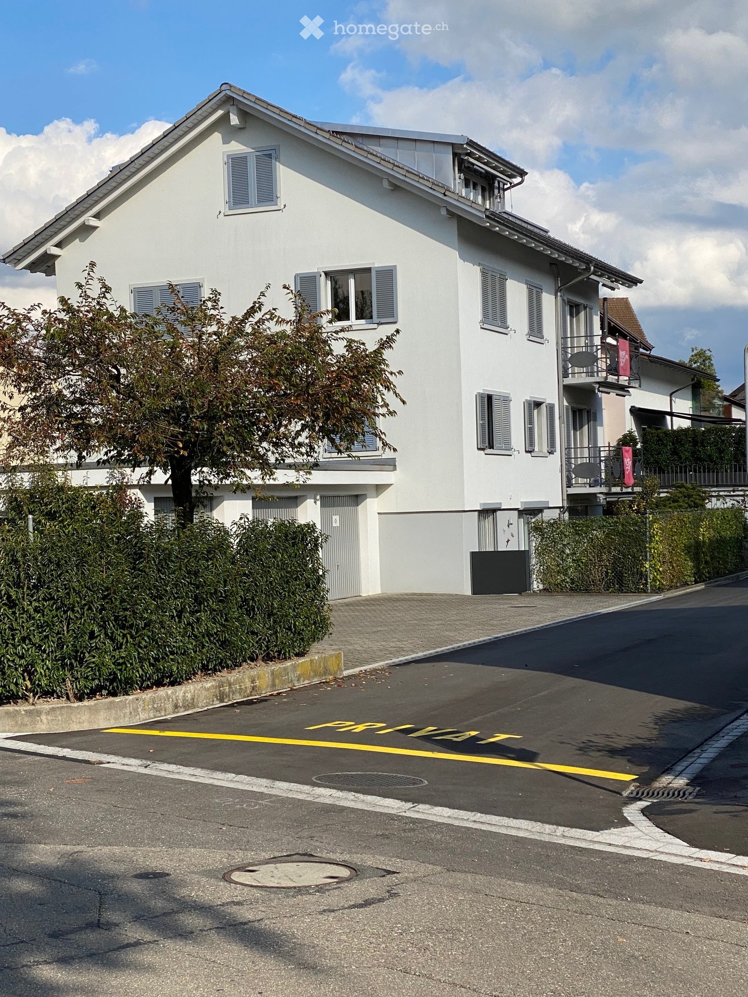 Two-story white house with multiple windows, shutters, and a small balcony on the upper floor. The house is surrounded by greenery and has a driveway marked with yellow lines.