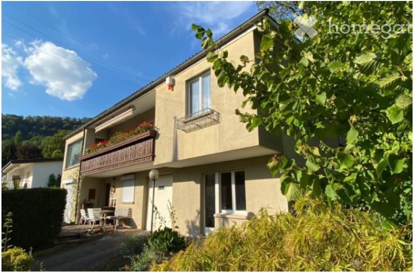 Two-story house with beige exterior, multiple windows, and a balcony with flower boxes. The house is surrounded by greenery and has a small patio area with outdoor furniture.