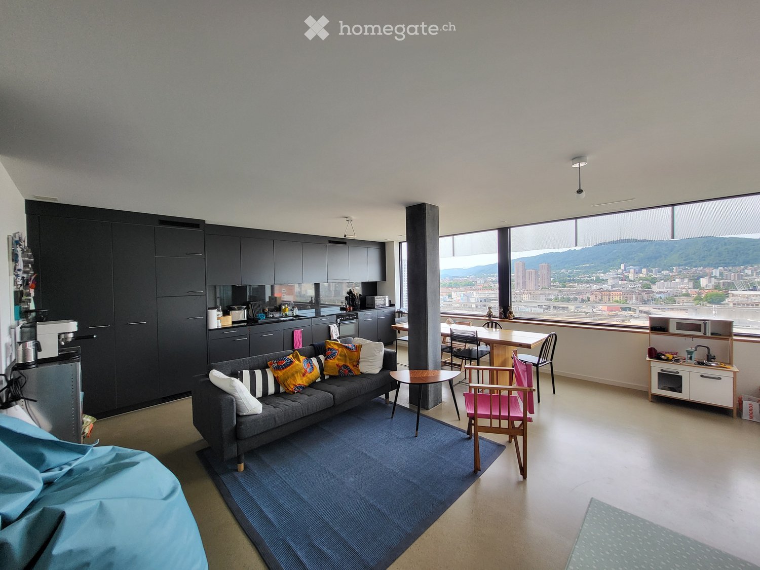 Modern, open-concept living space with a grey sofa, colorful cushions, coffee table, dining area with chairs and table, and a fully equipped kitchen with dark cabinets and stainless steel appliances.