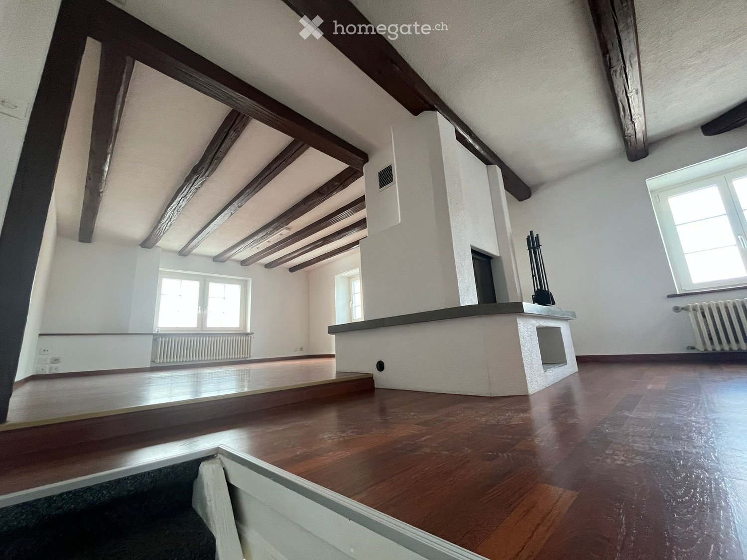 An empty room with wooden floors, exposed wooden beams on the ceiling, white walls, and large windows allowing natural light. There is a fireplace with a black mantelpiece and radiators along the walls.