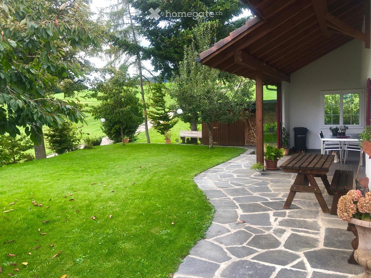 A well-maintained garden with a stone pathway leading to a wooden bench and a small seating area near the house. The area includes a grassy lawn, potted plants, and a view of a rural landscape with trees and hills.
