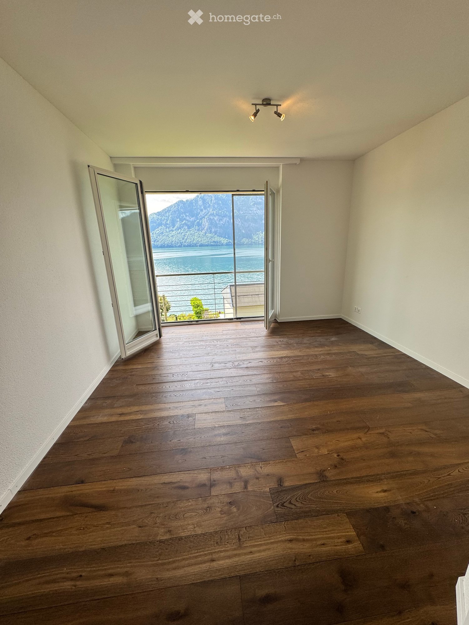 Empty room with wooden flooring, large sliding glass doors leading to a balcony with a scenic lake and mountain view.