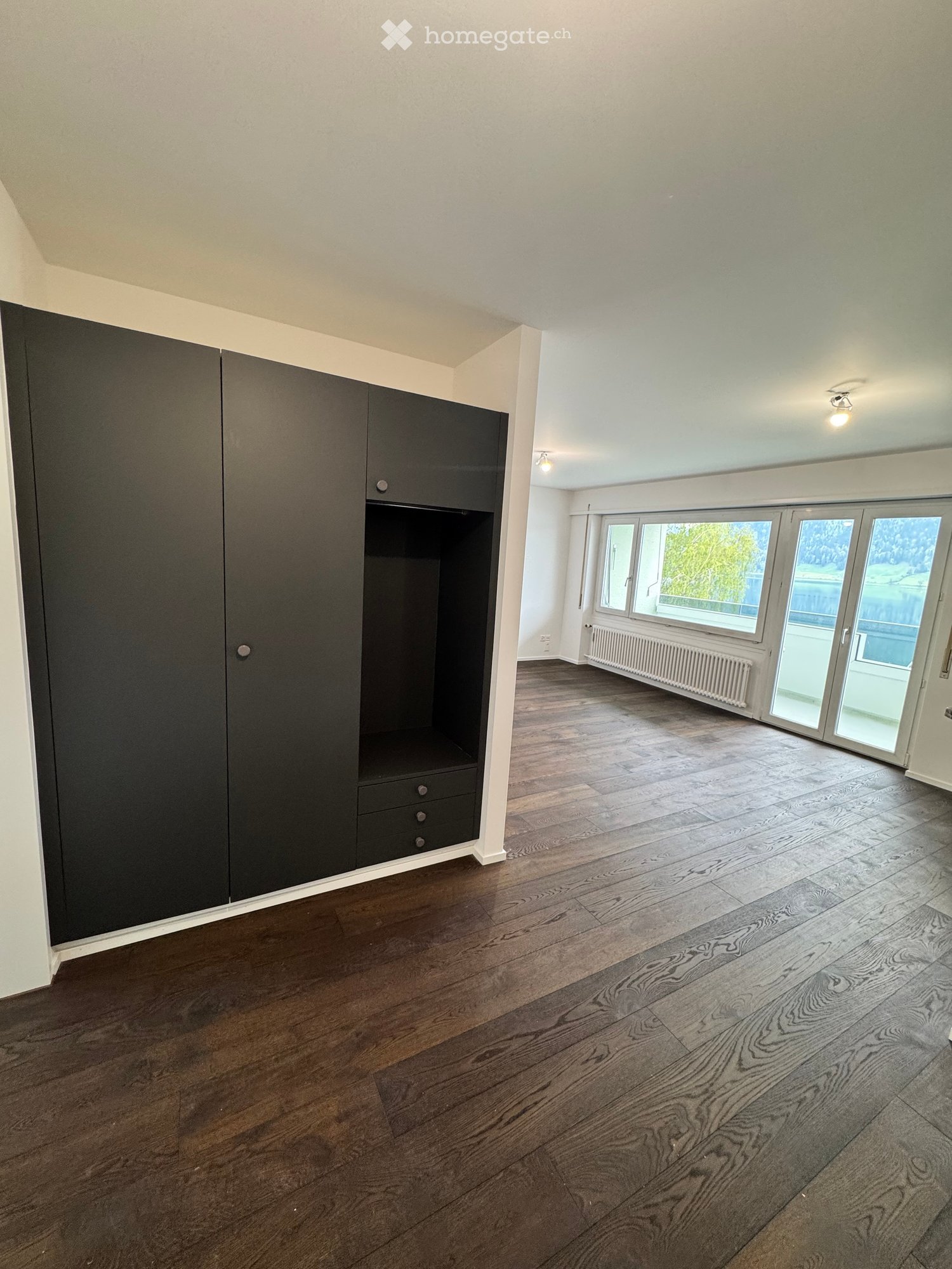 Empty room with wooden flooring, large windows, and a built-in wardrobe with drawers.