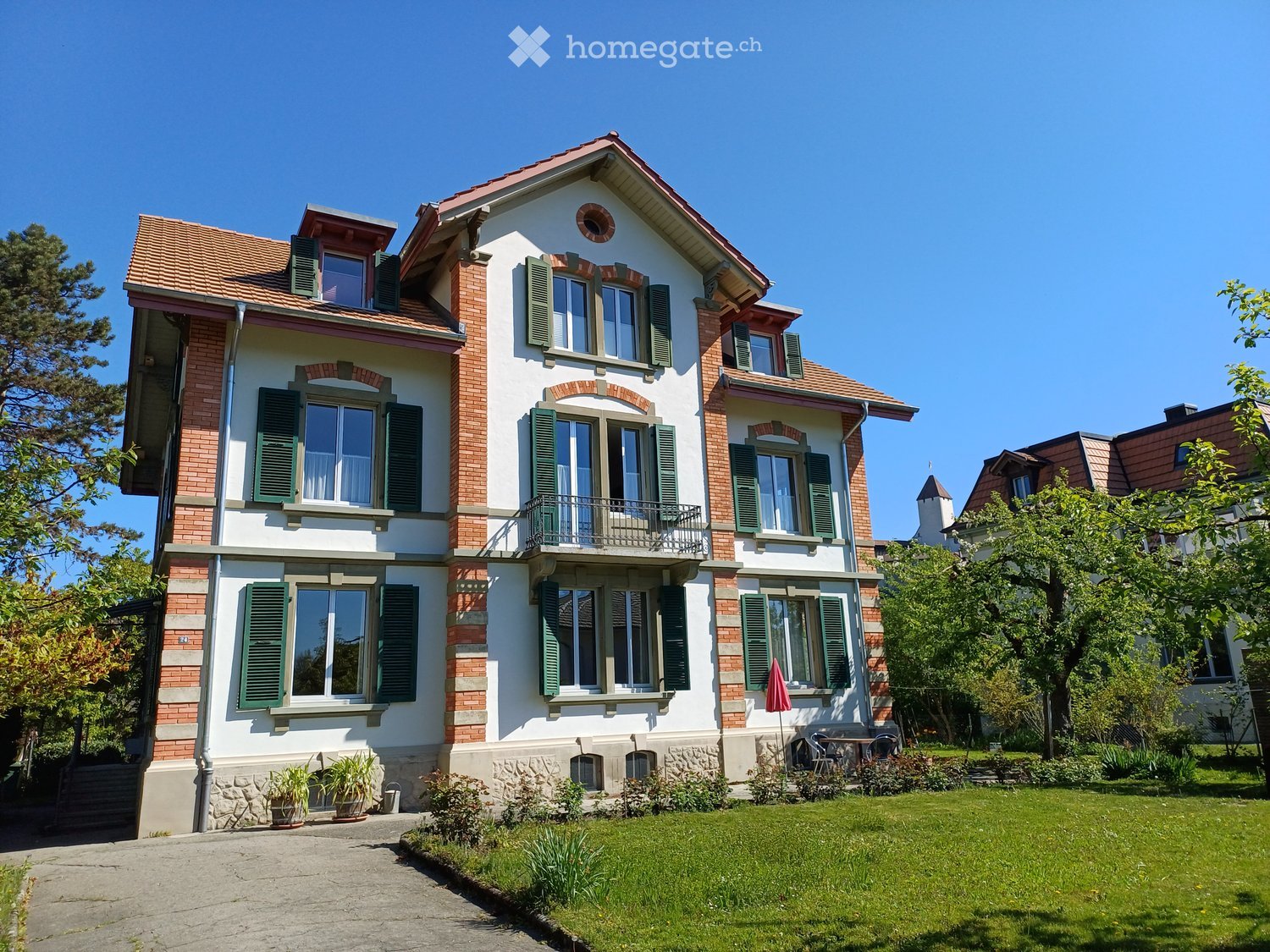 Two-story house with red-tiled roof, white and brick exterior, green shutters, multiple windows, and a small balcony. The house is surrounded by a well-maintained lawn and garden with trees and shrubs.