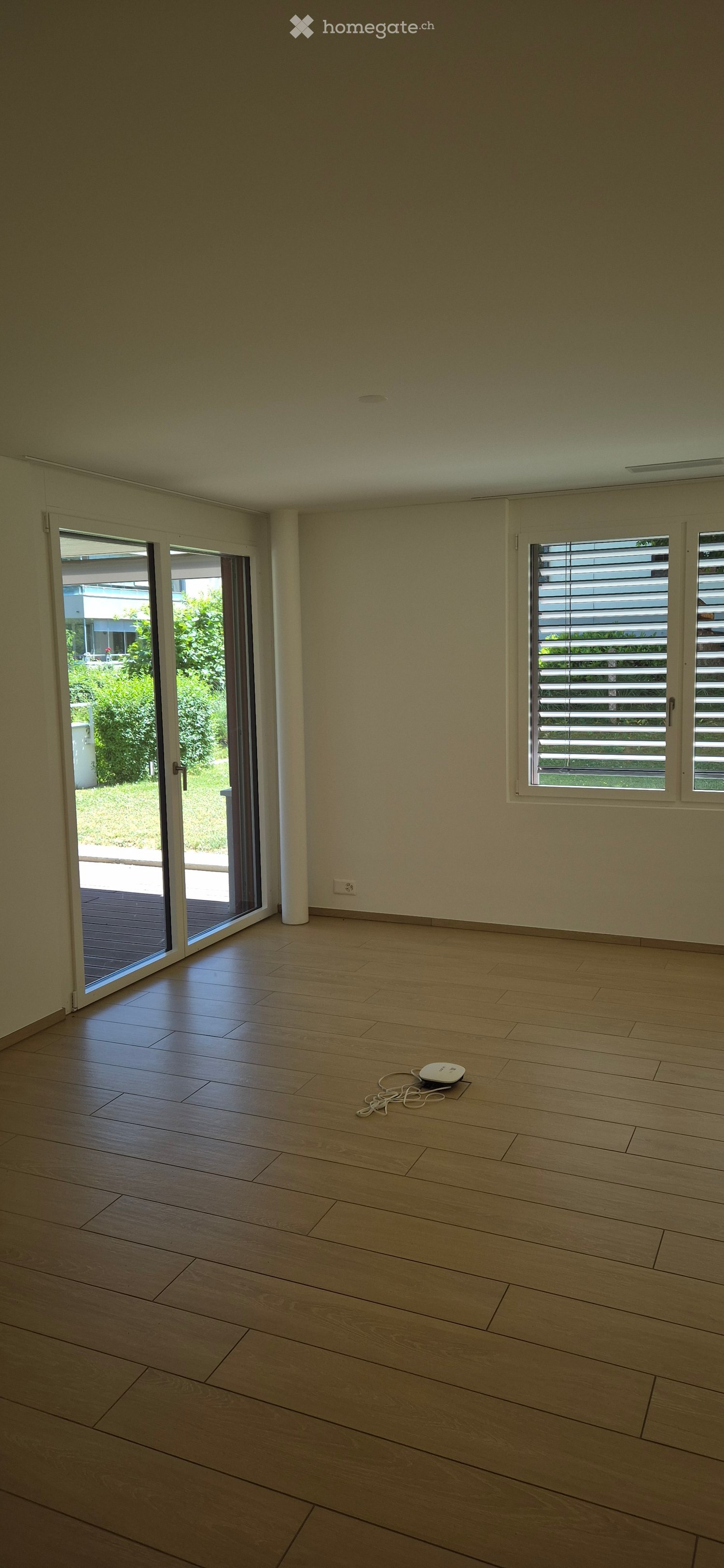 Empty room with light wooden flooring, white walls, large windows with blinds, and a sliding glass door leading to an outdoor area.
