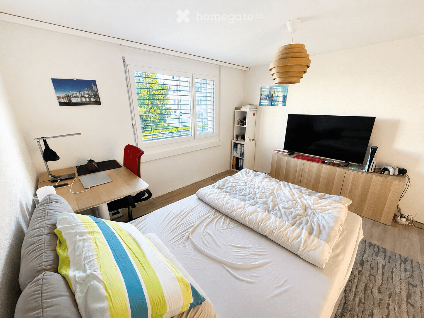 A modern bedroom with a double bed, desk, chair, laptop, and a TV stand with a screen. The room features large windows with blinds, a hanging lamp, and a bookshelf.
