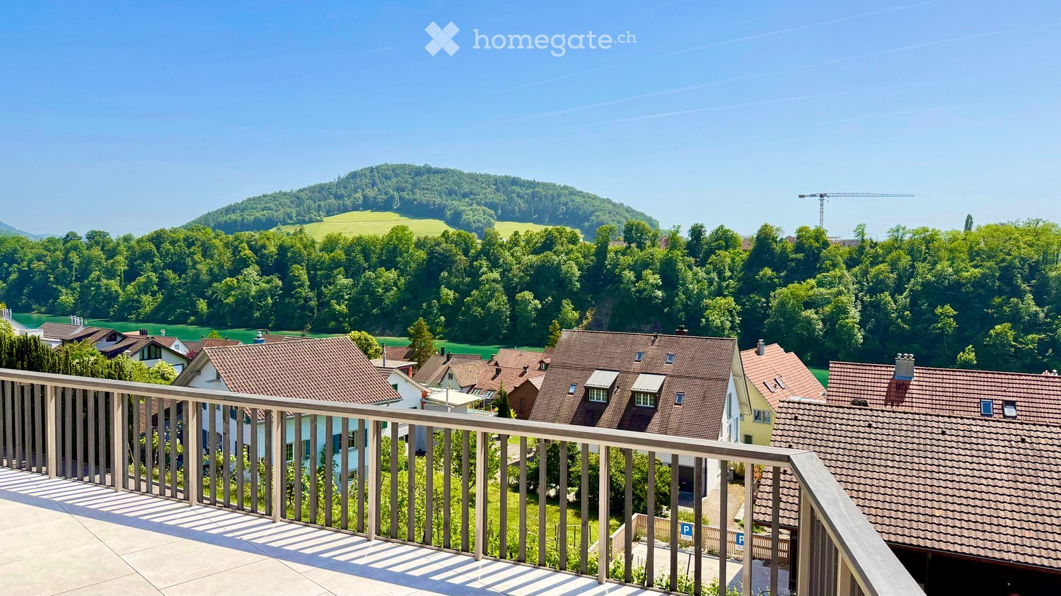 Balcony overlooking a scenic town with red-roofed houses, lush green trees, and distant hills under a clear blue sky.