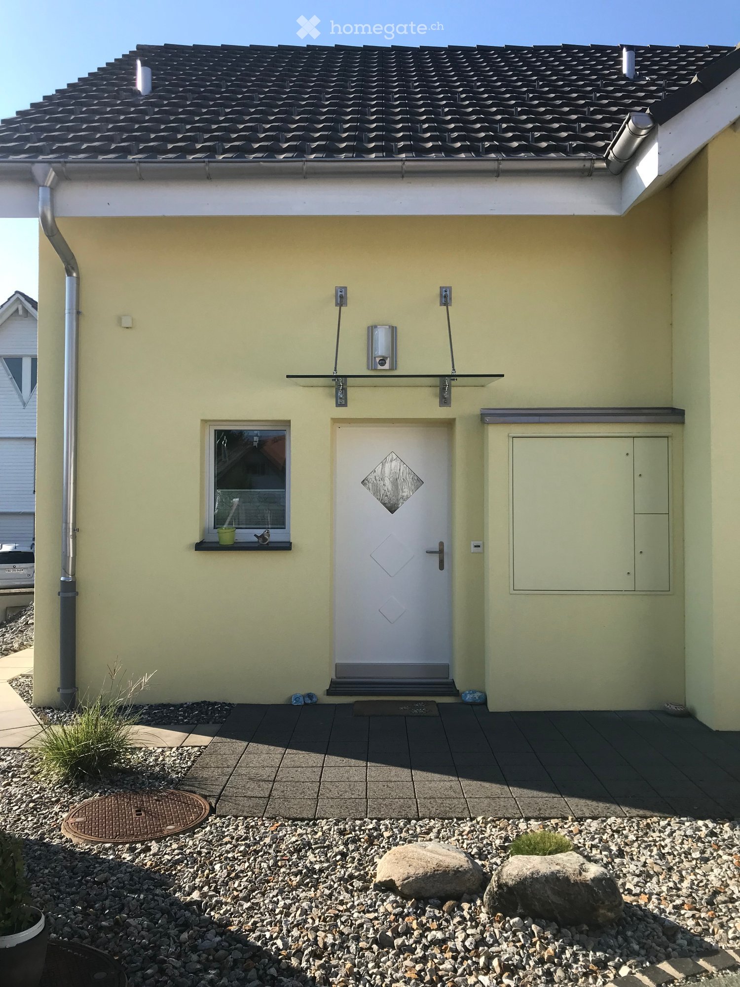 Single-story house with light yellow exterior, white front door, small window with a plant on the sill, and a tiled entrance area.
