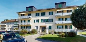 3-story apartment building with balconies, white and yellow exterior, surrounded by landscaped grounds, parking spaces in front