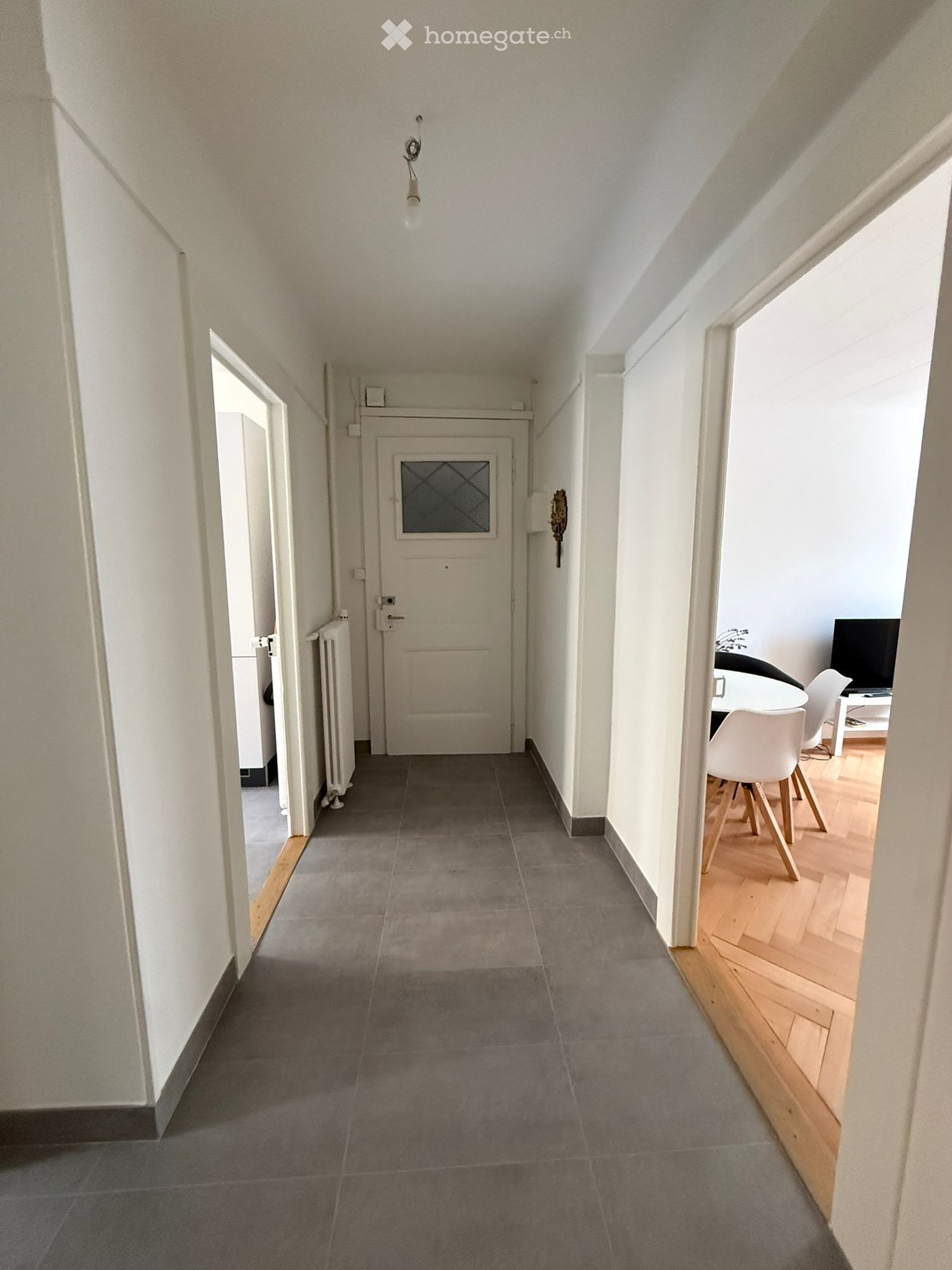A narrow hallway with grey tiled flooring, white walls, and a ceiling light fixture. The hallway leads to a door with a glass panel and a radiator on the left side. To the right, a glimpse of a dining area with white chairs and a table is visible.