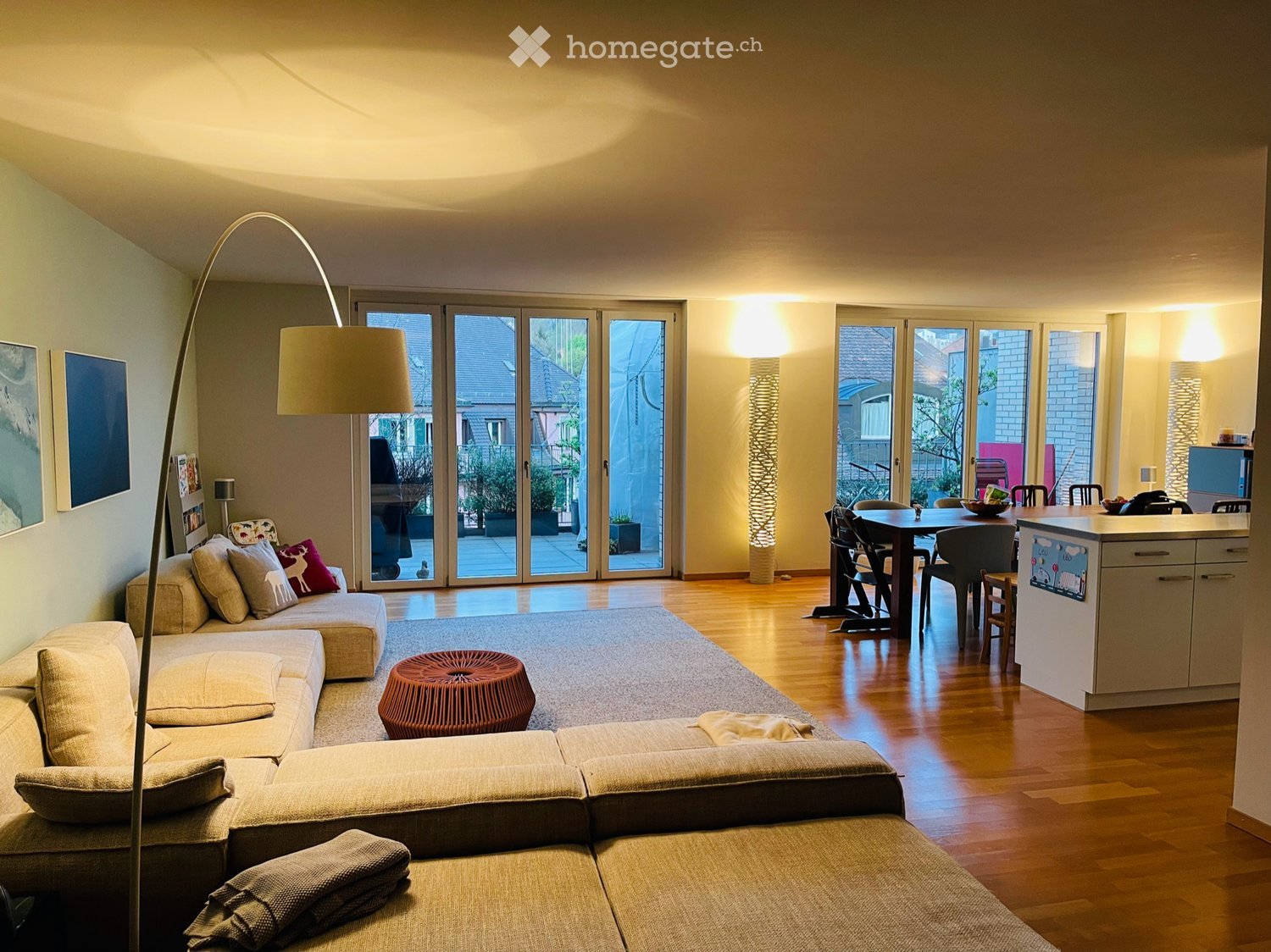 Open-plan living and dining area with a sectional sofa, a round wooden stool, a dining table with chairs, a modern kitchen island, large sliding glass doors leading to a balcony, and a floor lamp.