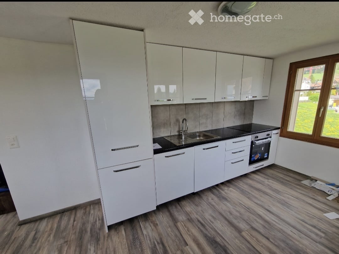 Modern kitchen with white cabinets, black countertops, a sink, and an oven. The floor is made of wood, and there is a window with a view of greenery.
