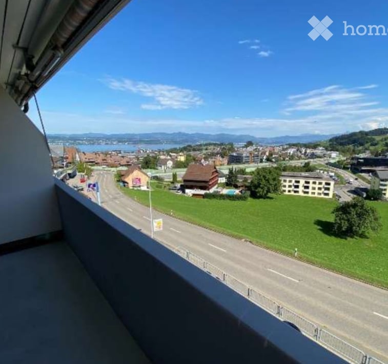 Balcony with a scenic view of a town, green fields, and distant mountains under a clear blue sky.