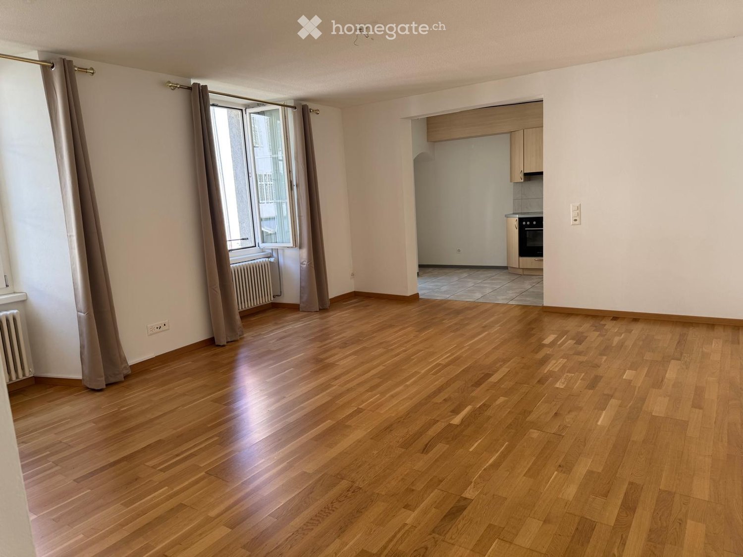 Empty room with wooden flooring, large window with curtains, radiator, and an adjacent kitchen area with cabinets and appliances.