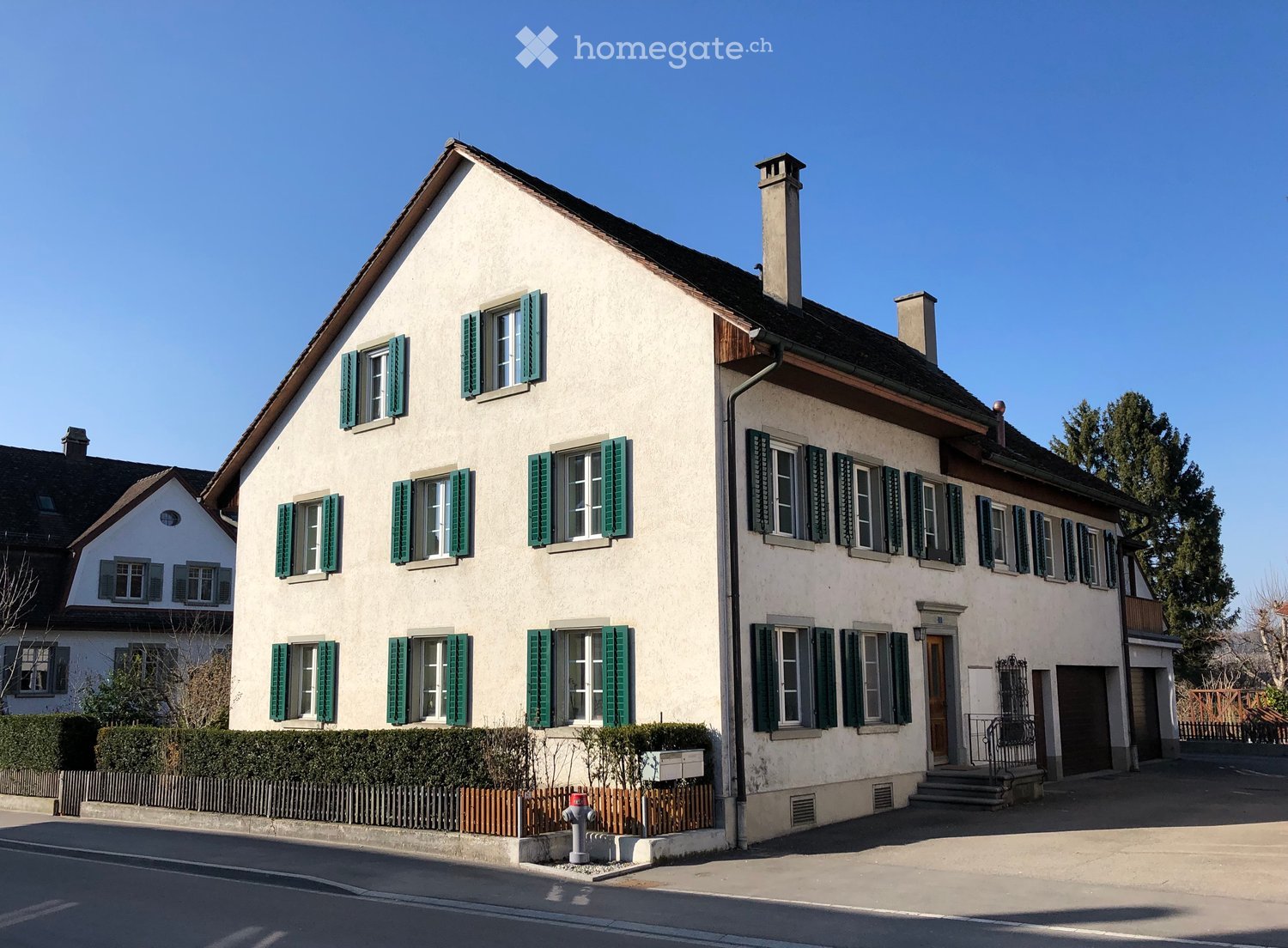 Two-story house with white walls, green shutters, and multiple windows. Features a chimney, a small garden with a fence, and a front staircase leading to the entrance.