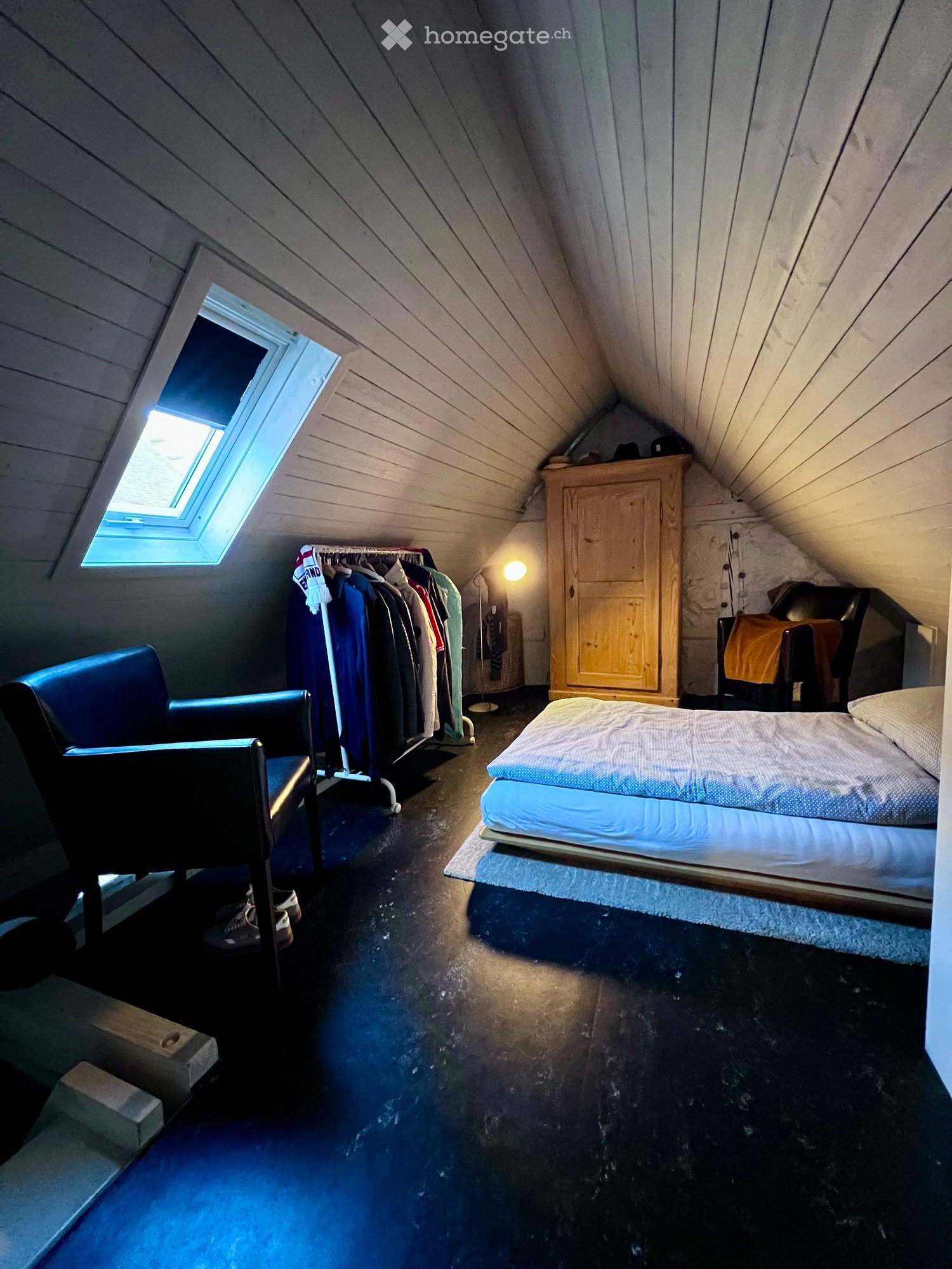 Attic bedroom with slanted wooden ceilings, skylight window, bed, chair, wardrobe, and clothing rack.
