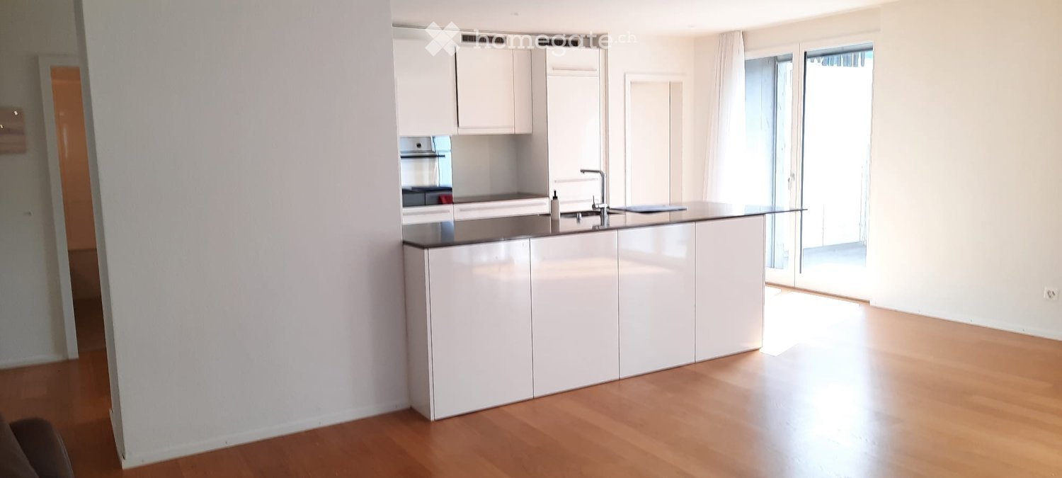 Modern kitchen with white cabinets, a central island, and a sink. The space features wooden flooring and large windows allowing natural light to fill the room.