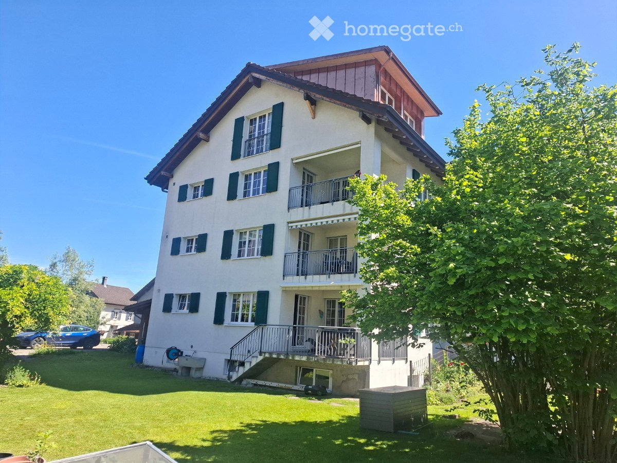 Two-story house with white walls, green shutters, multiple windows, and balconies. The house has a well-maintained lawn and is surrounded by trees.