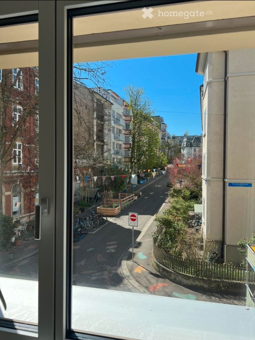 View from a balcony overlooking a quiet urban street lined with buildings, bicycles, and small garden areas.